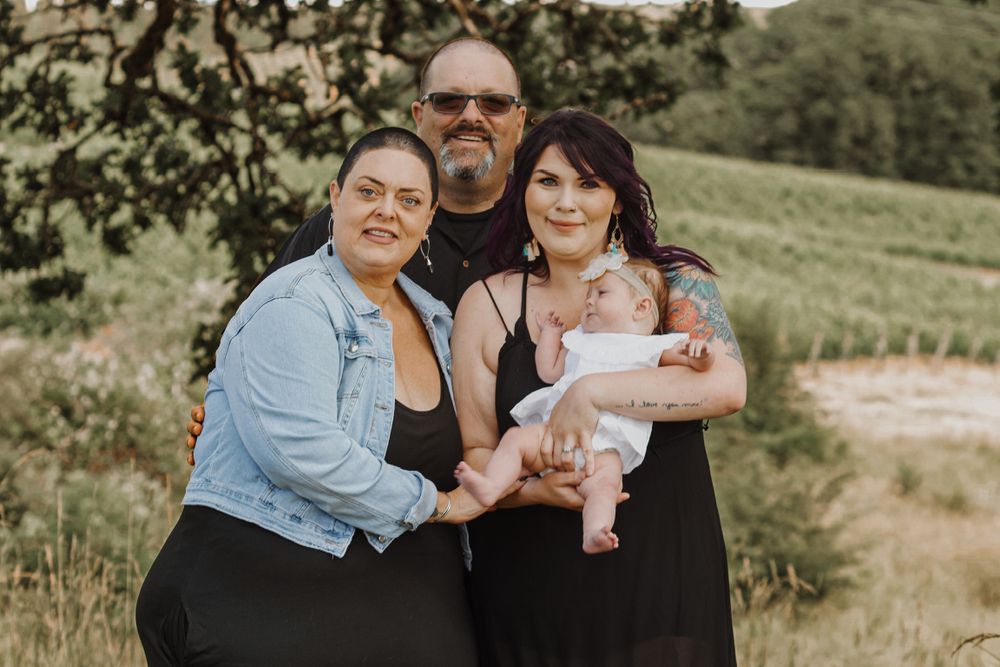 A family of three adults proudly holds a newborn baby in an outdoor field setting.