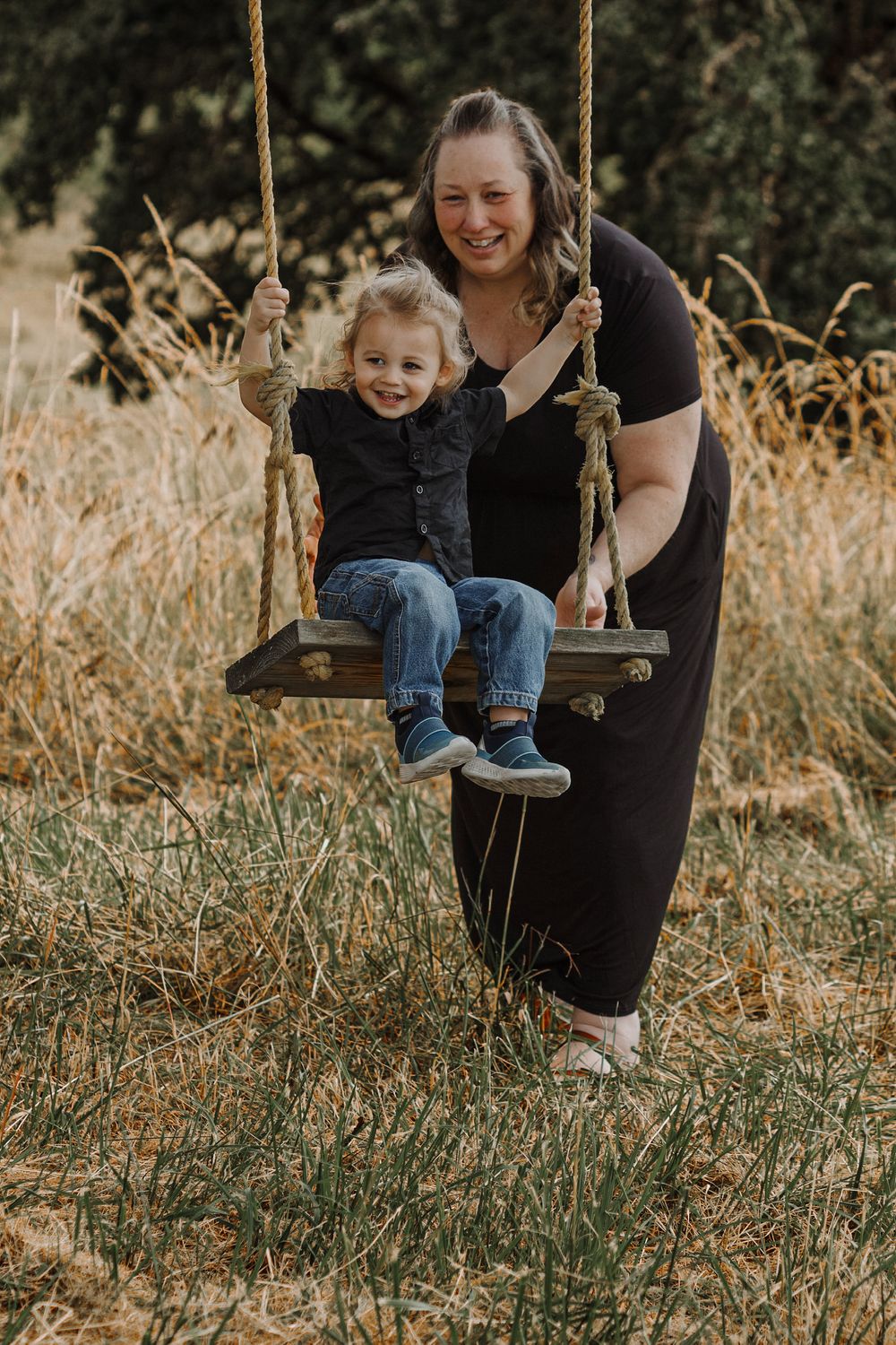 A laughing mother pushes her toddler on a rustic rope swing outdoors in a golden field.