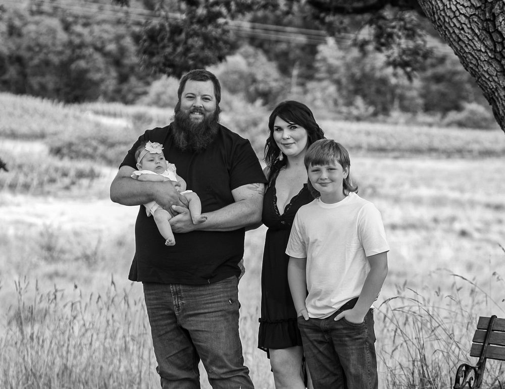 Black and white family portrait of bearded father holding newborn with mother and son outdoors.