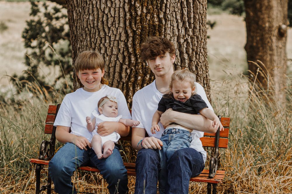 Two teenage boys sit on a bench under a tree each holding a baby in a sunny field.