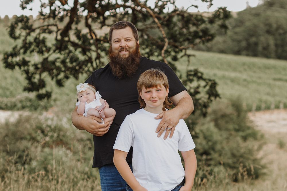 Bearded father holds newborn baby while standing with young son in a lush green outdoor setting.
