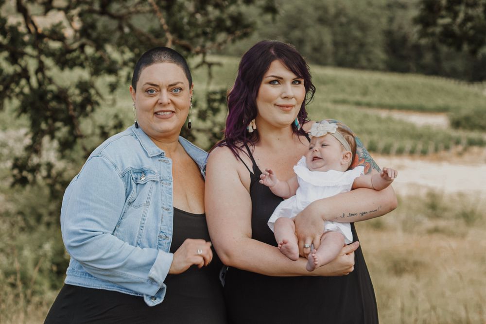 Two women stand outdoors in nature, one holding a sleeping newborn baby in a black dress.