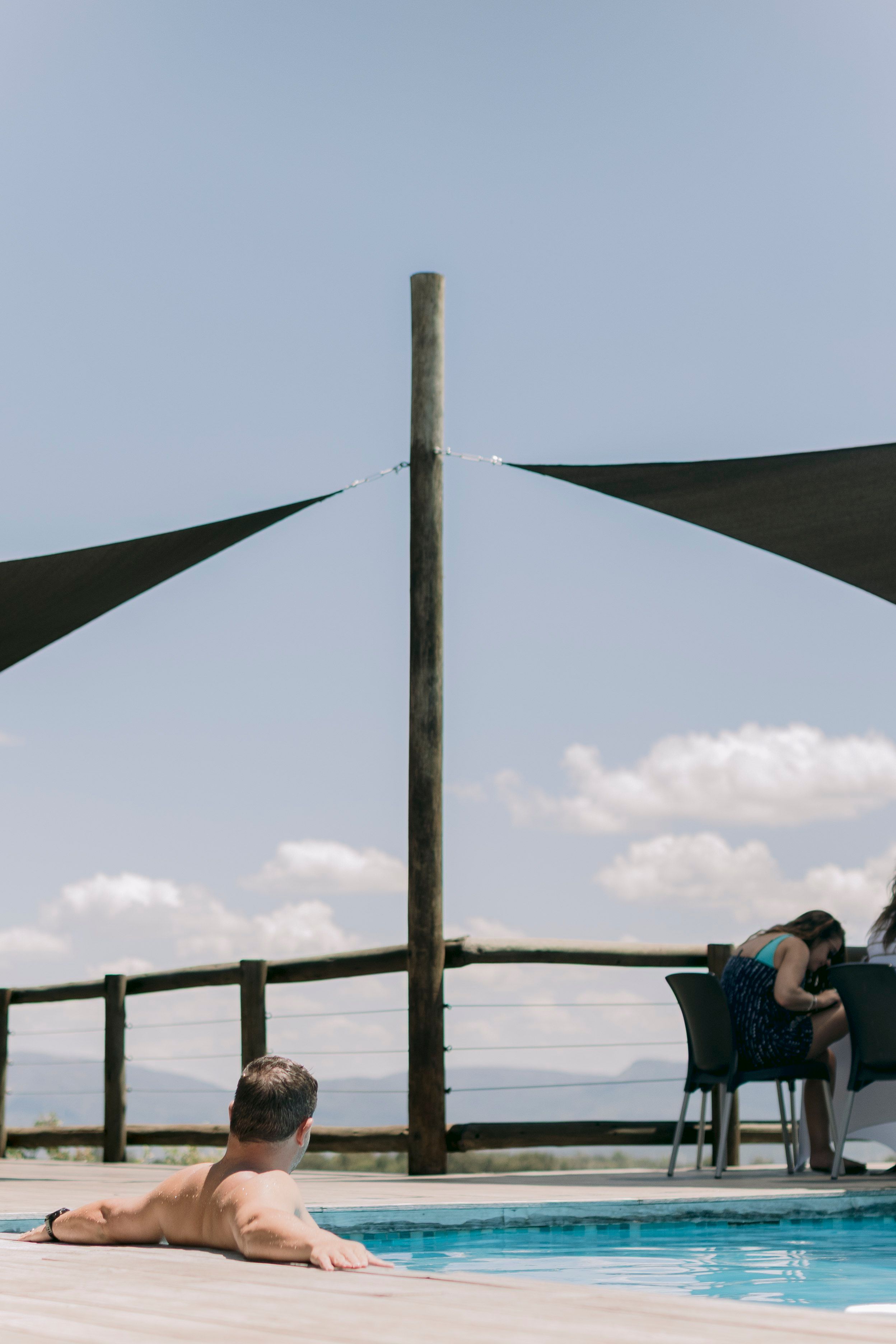 Man relaxing at a luxury safari lodge infinity pool with shade sails and scenic mountain views.