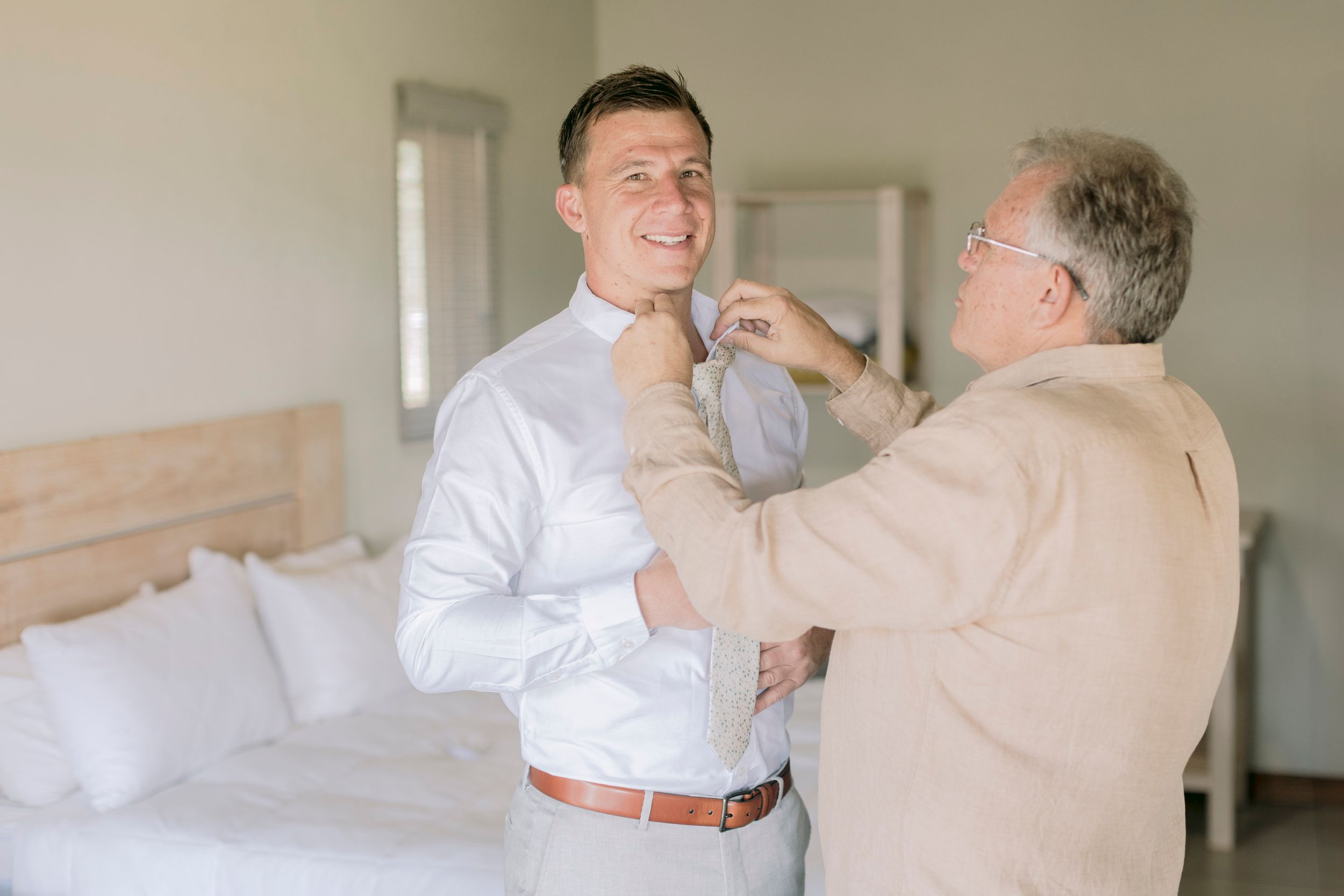 Smiling groom getting his tie adjusted by his father in a bright white wedding suite bedroom.