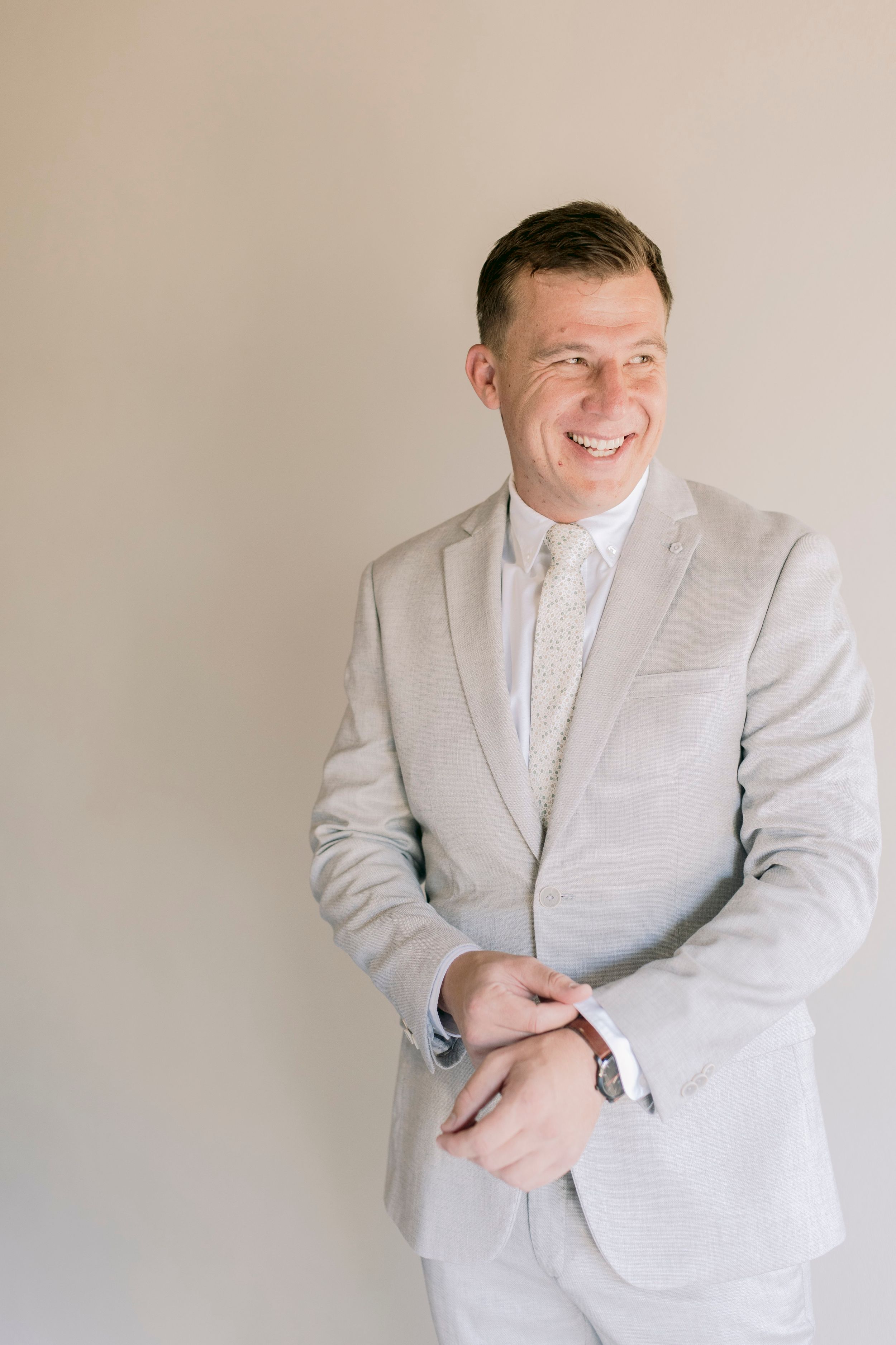 Smiling groom in light gray suit and cream tie adjusting cufflinks before wedding.