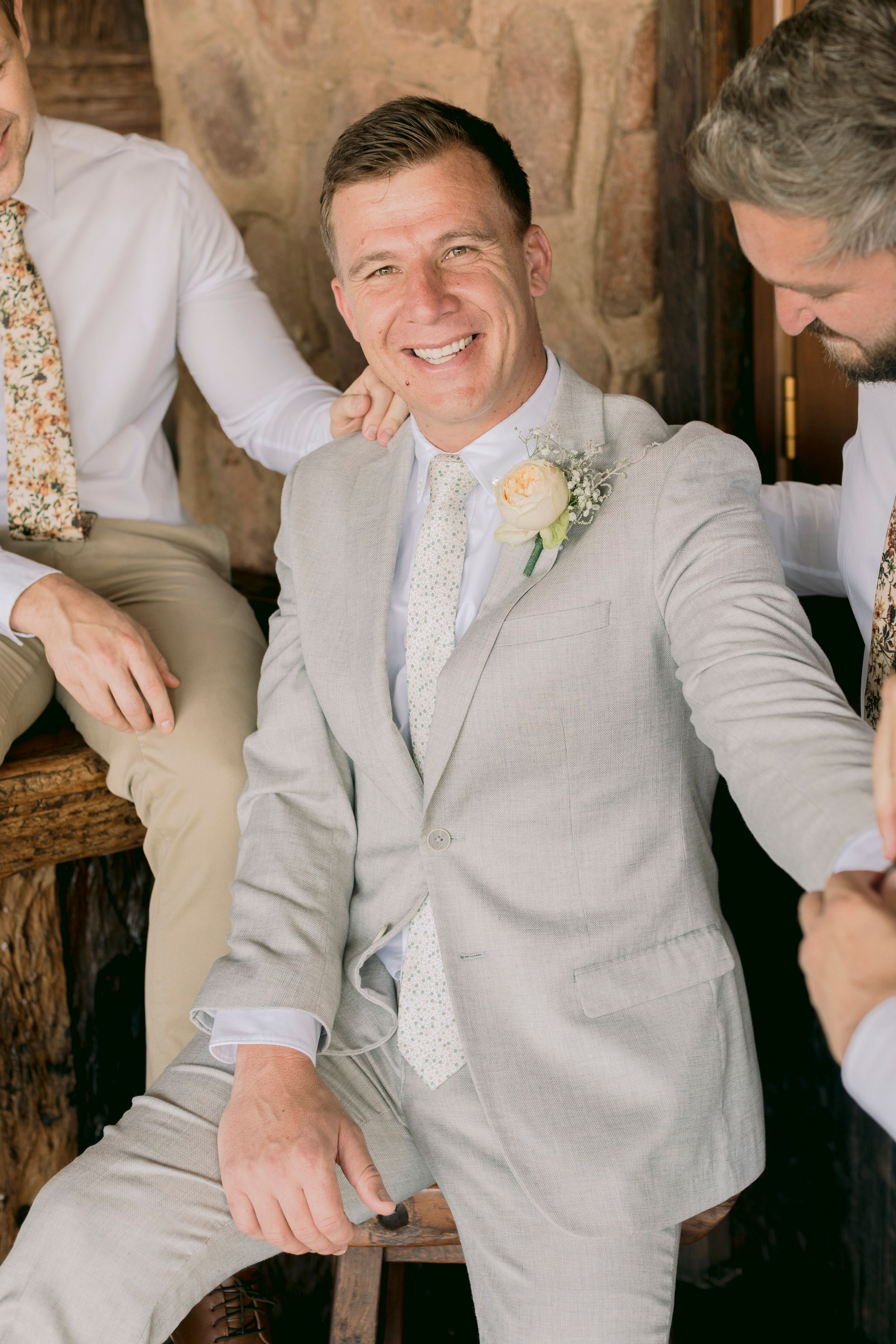 Smiling groom in light grey suit with floral boutonniere sitting with groomsmen at rustic wedding venue.