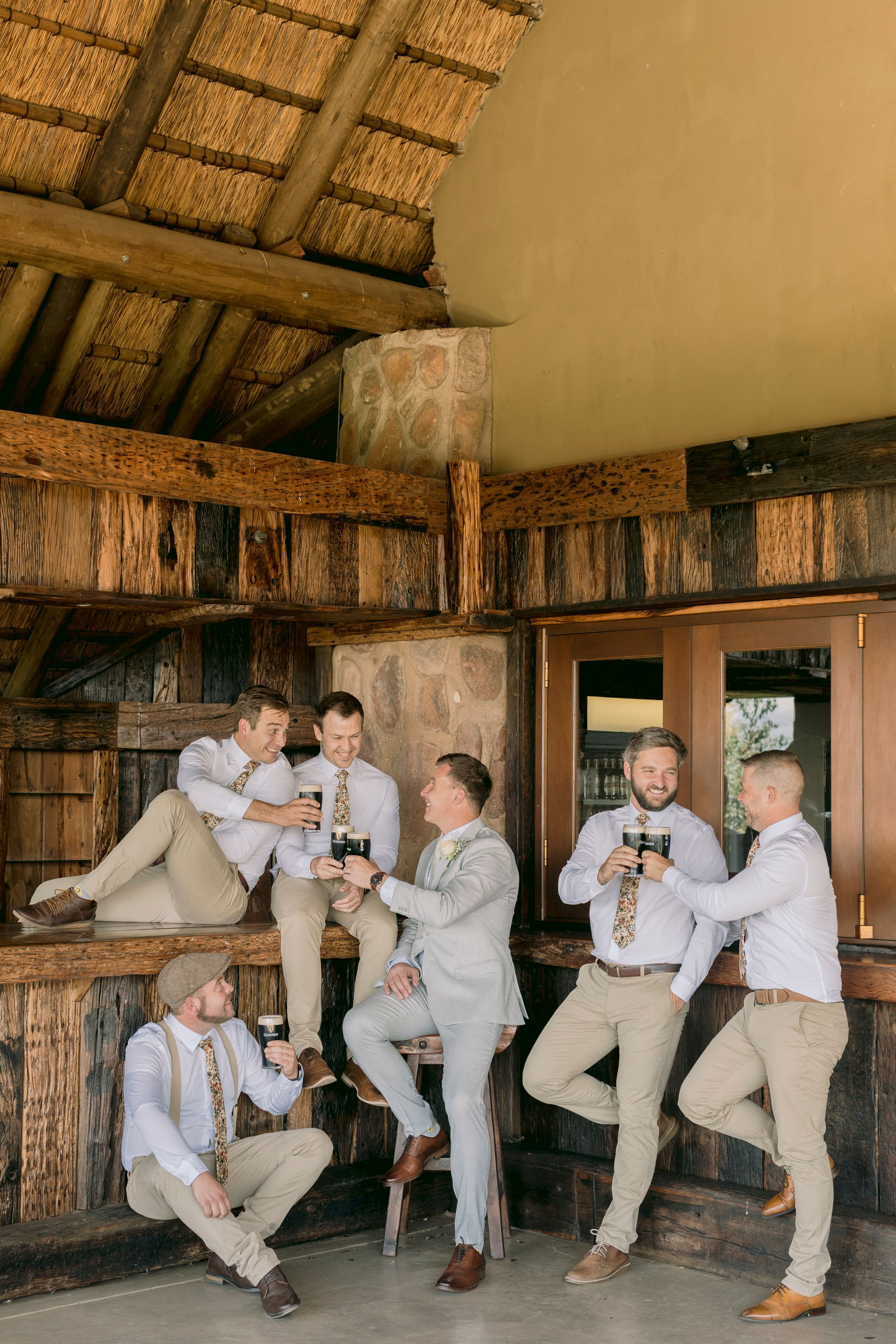 Groom and groomsmen toasting with dark beer bottles inside rustic thatched-roof wedding venue bar.