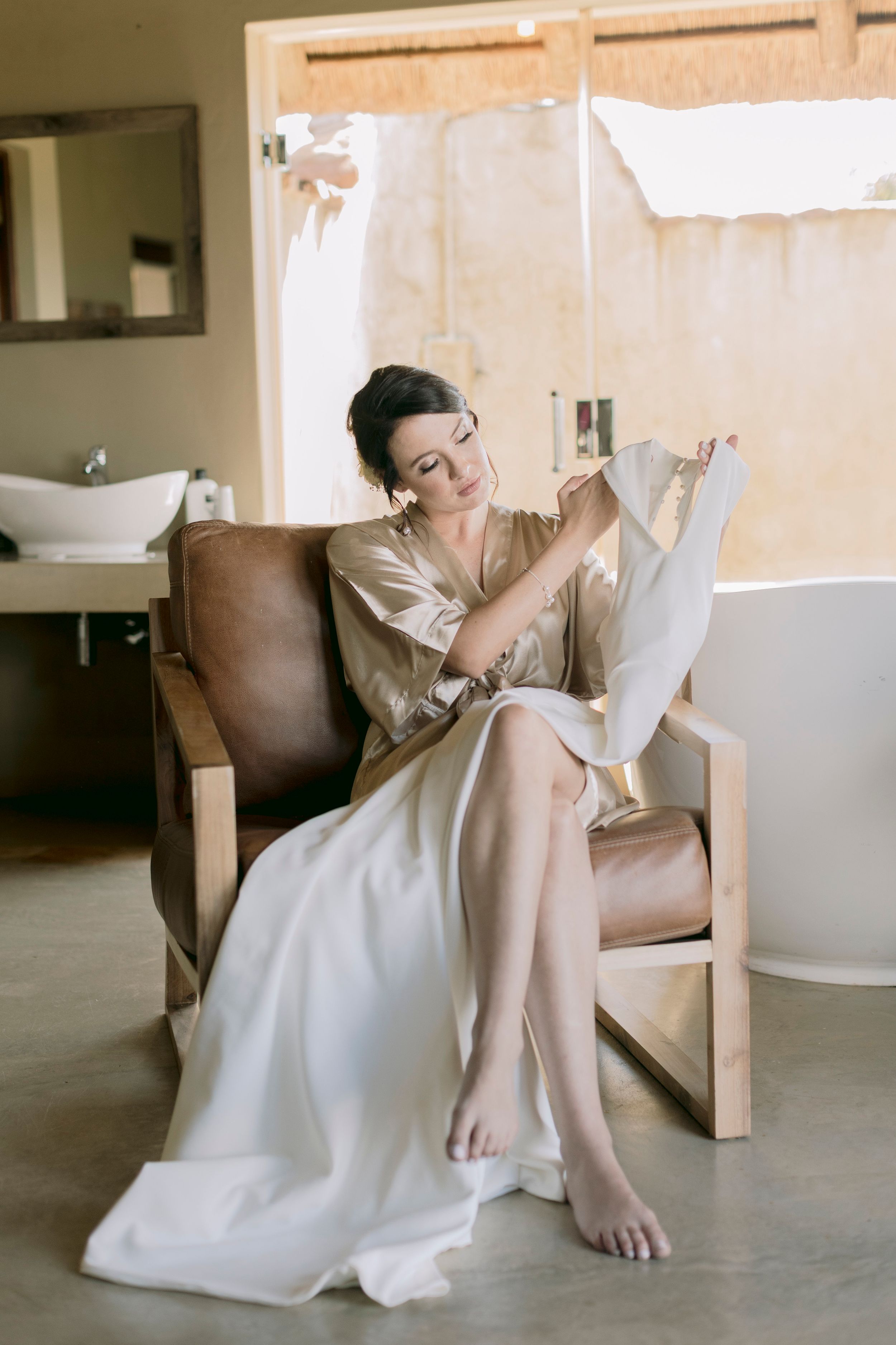 Bride in gold satin robe reading a note while seated in leather chair during wedding preparations.