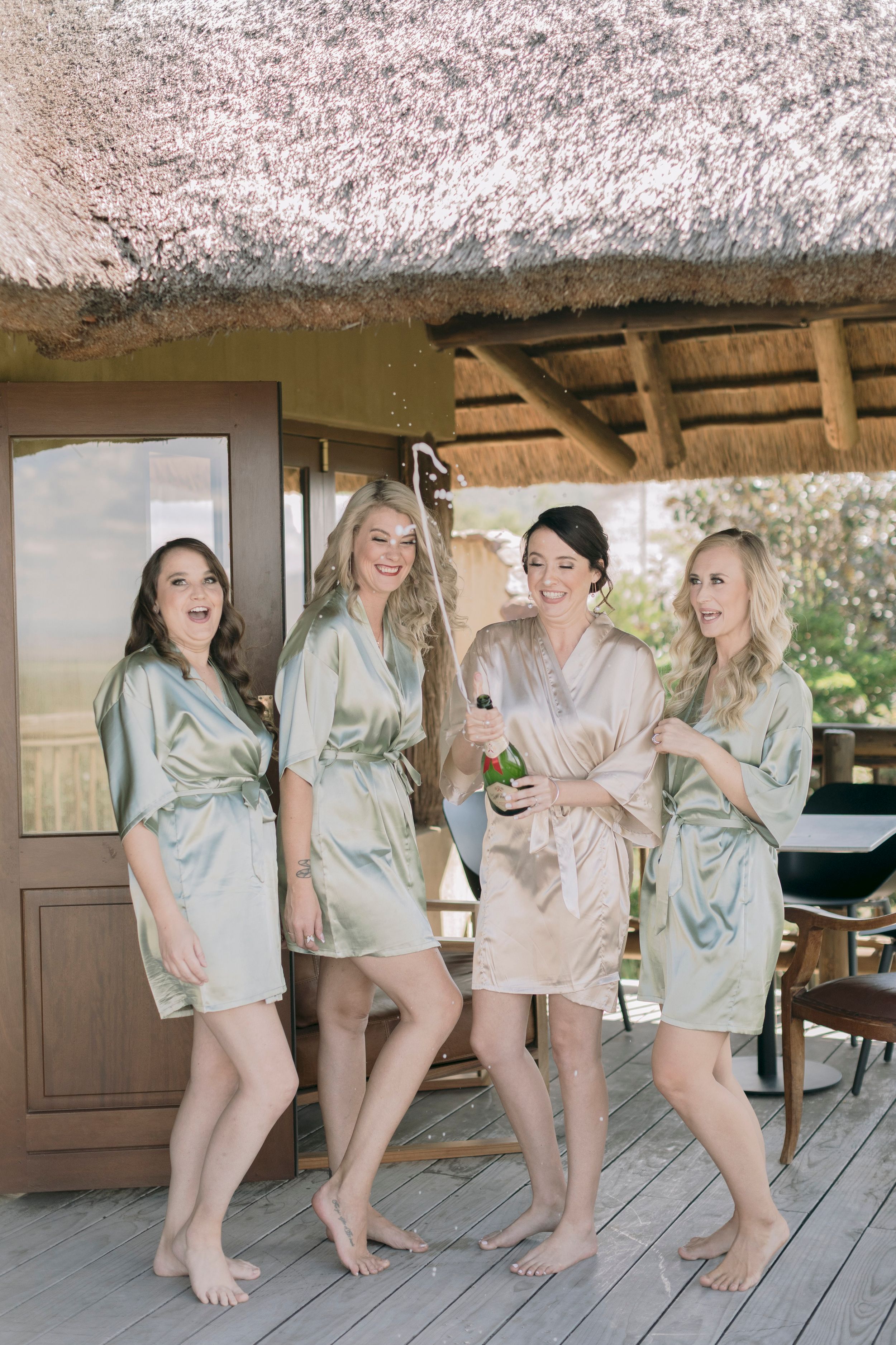 Bride and bridesmaids in satin robes celebrating with champagne on wooden deck at safari wedding venue.