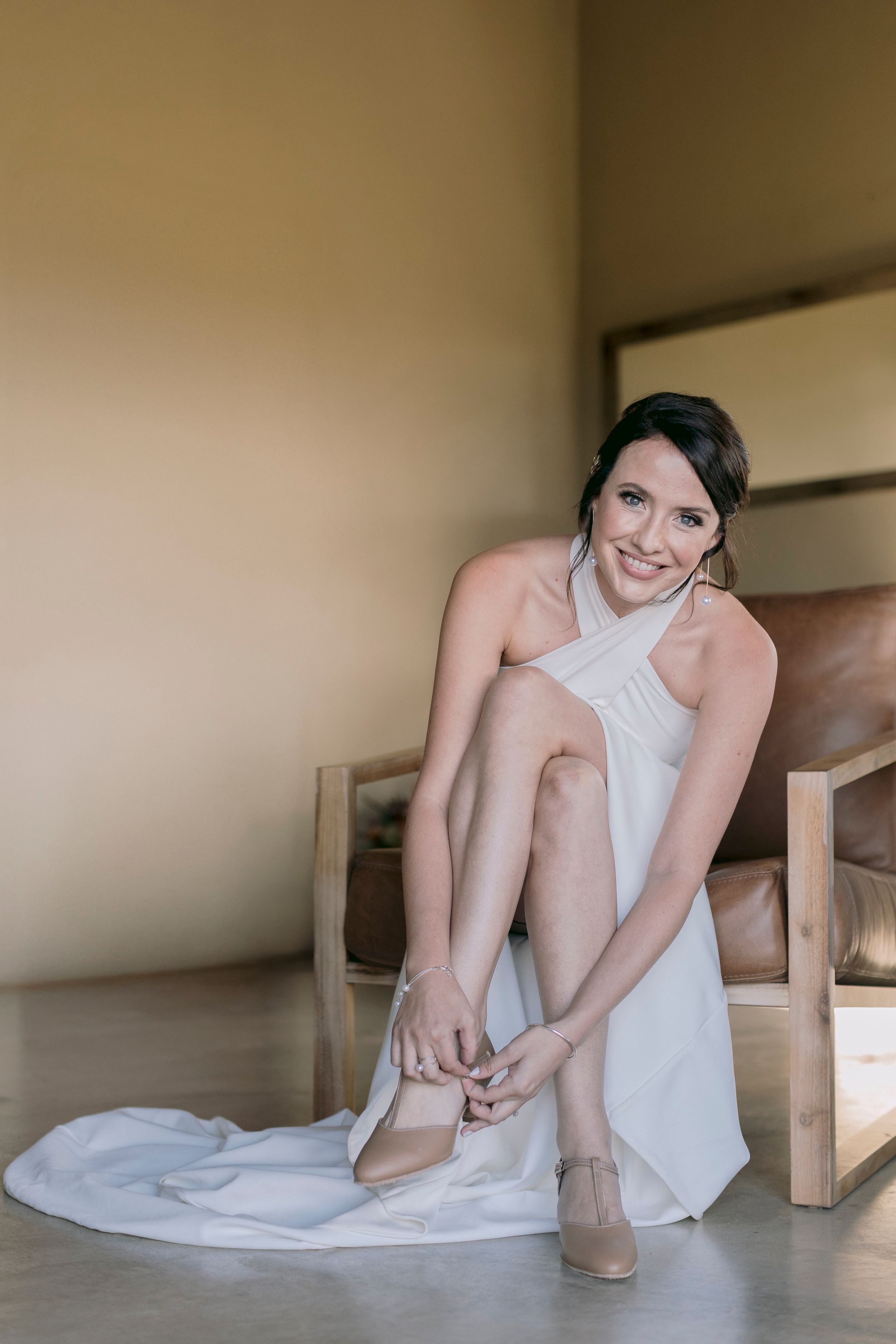 Bride in white halter wedding dress putting on nude heels while seated in leather chair.