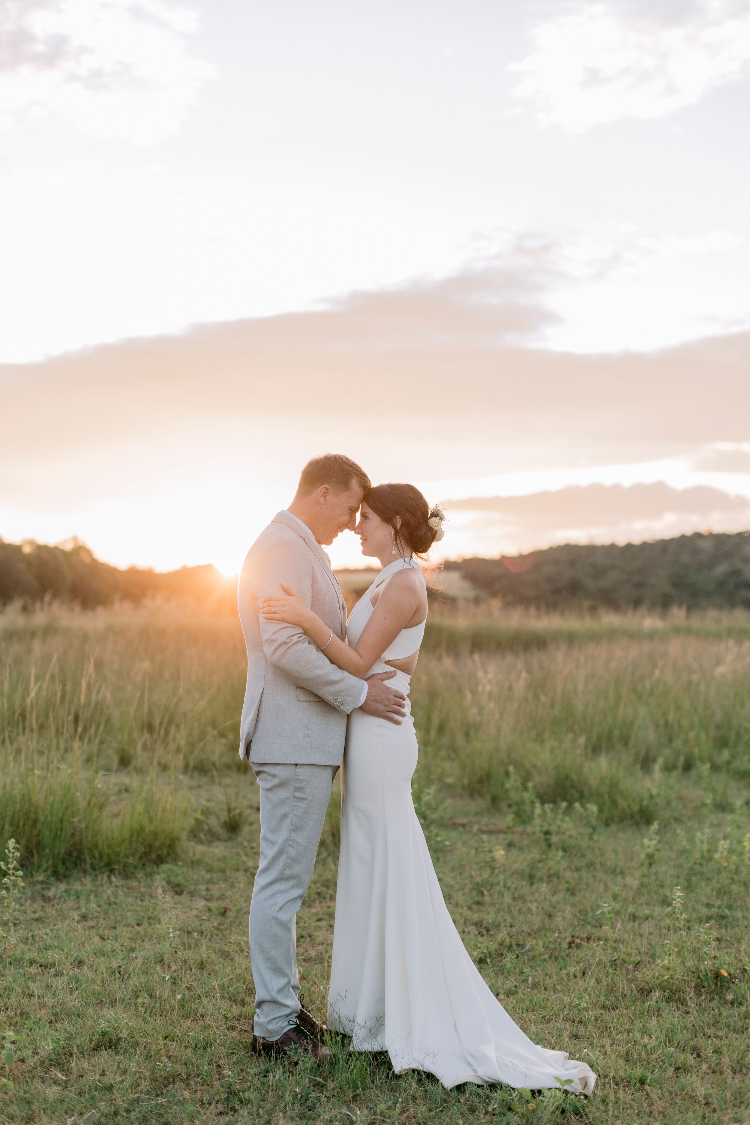 Newlyweds share an intimate forehead-to-forehead moment in an open field bathed in warm golden sunset light.