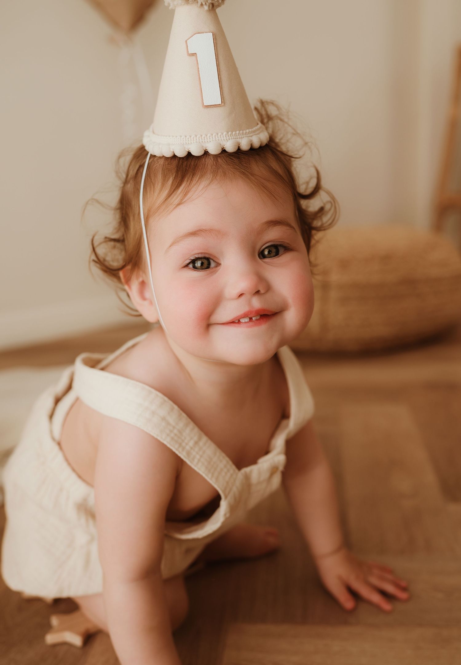 Small child in a beige dress and party hat sits on floor next to peach balloon during first birthday photoshoot.