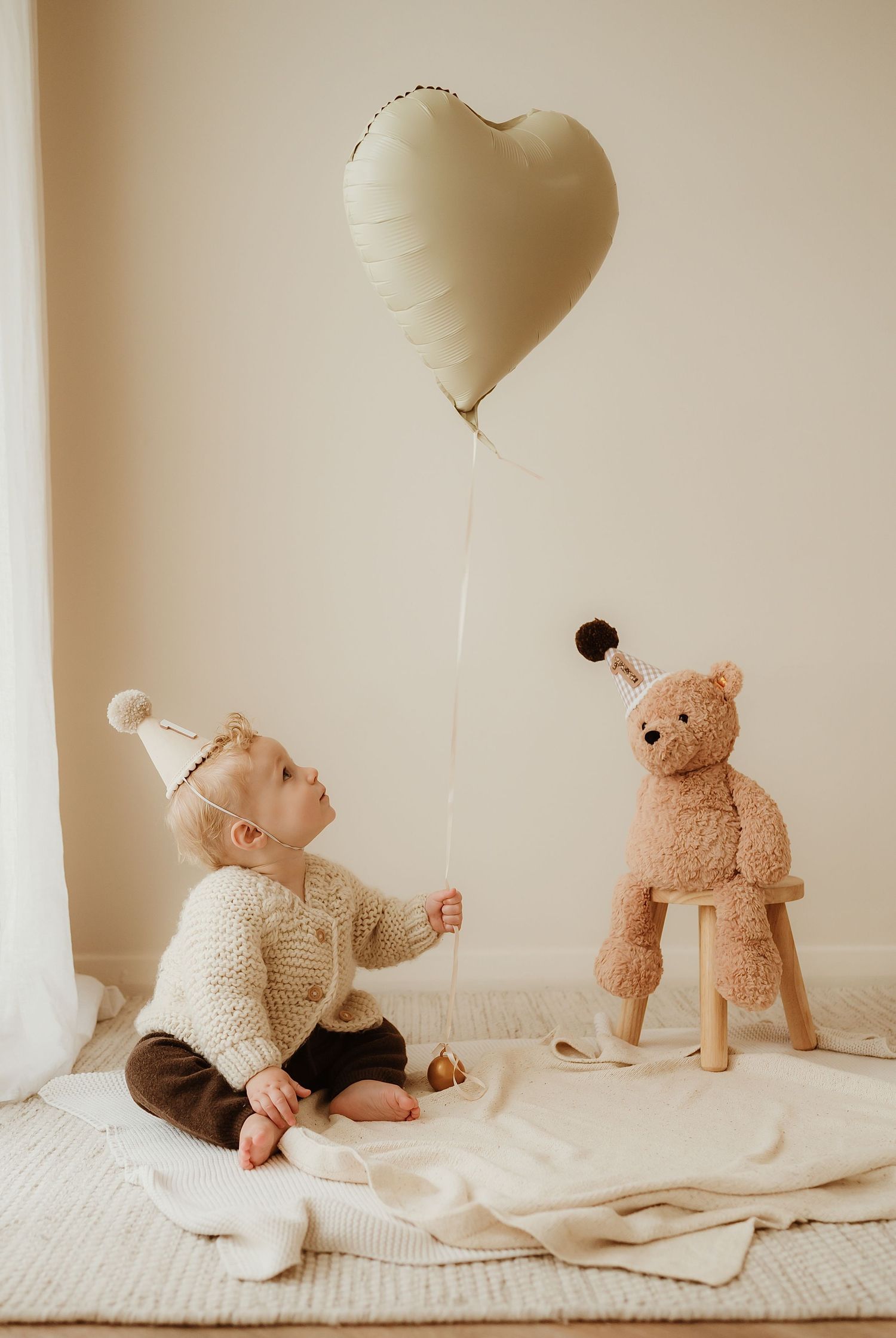 Small child in a beige dress and party hat sits on floor next to peach balloon during first birthday photoshoot.