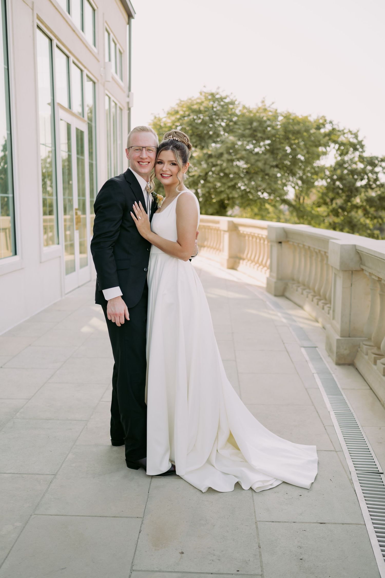 A couple in formal wedding attire embraces on an elegant stone balcony next to a glass building on their special day.