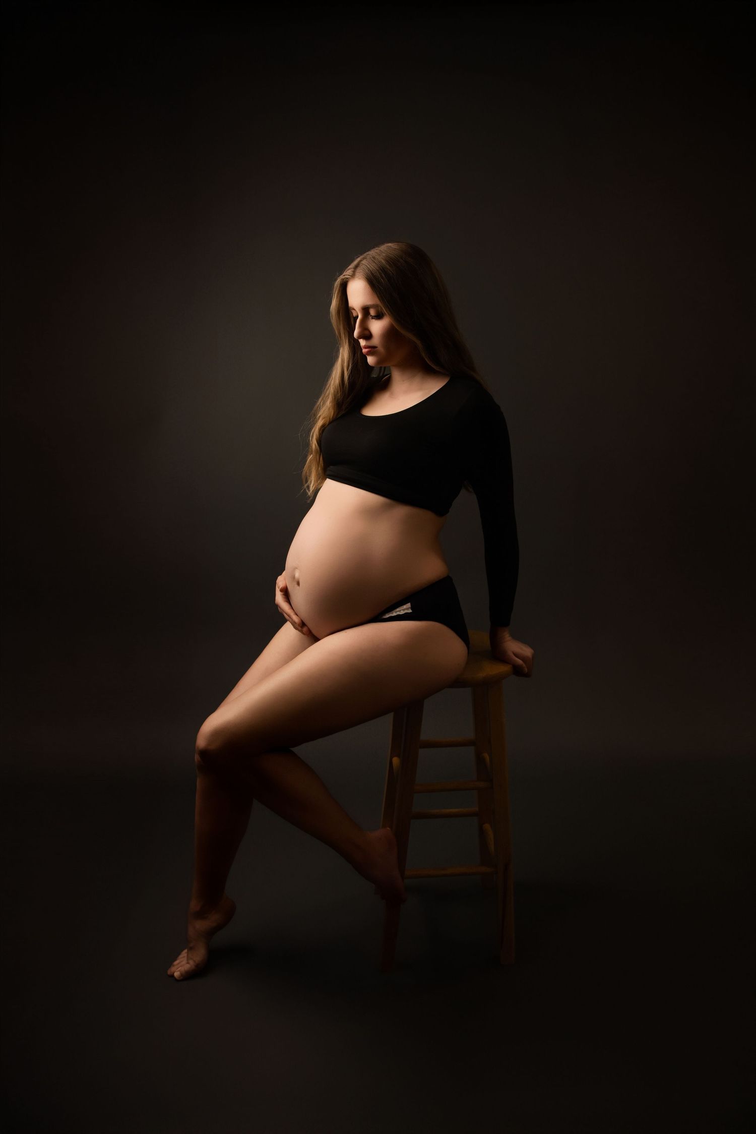 A dramatic maternity portrait shows an expectant mother in black attire seated on a wooden stool against a dark background.