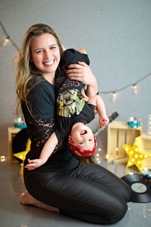 Parent in black attire playfully holds infant upside down against backdrop of decorative string lights and star decor.