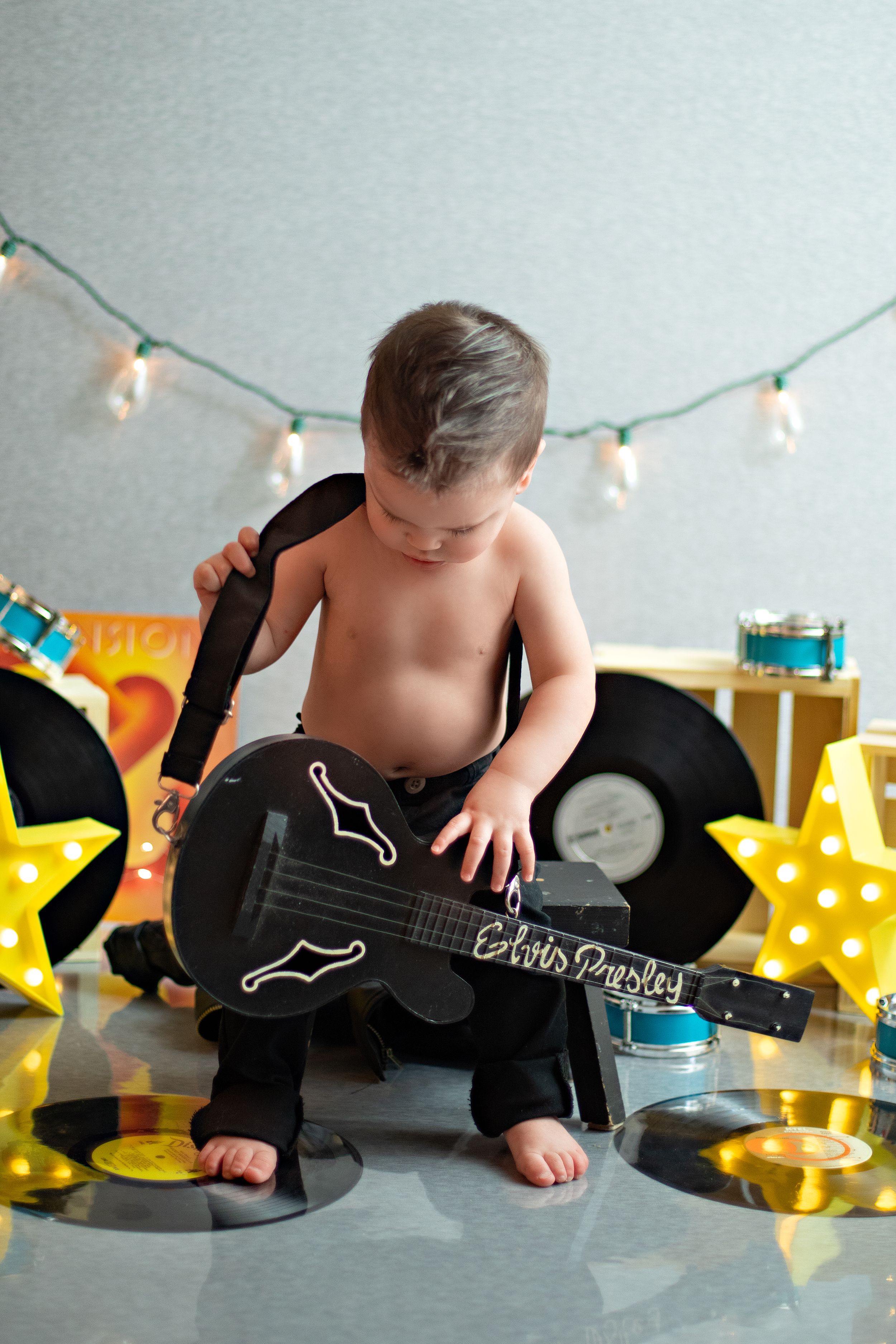A baby plays with a black guitar surrounded by vinyl records and star lights in a studio photo shoot setup.