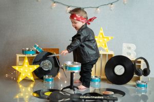 Young child in dark outfit and red headband stands among vinyl records, illuminated star lights and blue gift boxes.