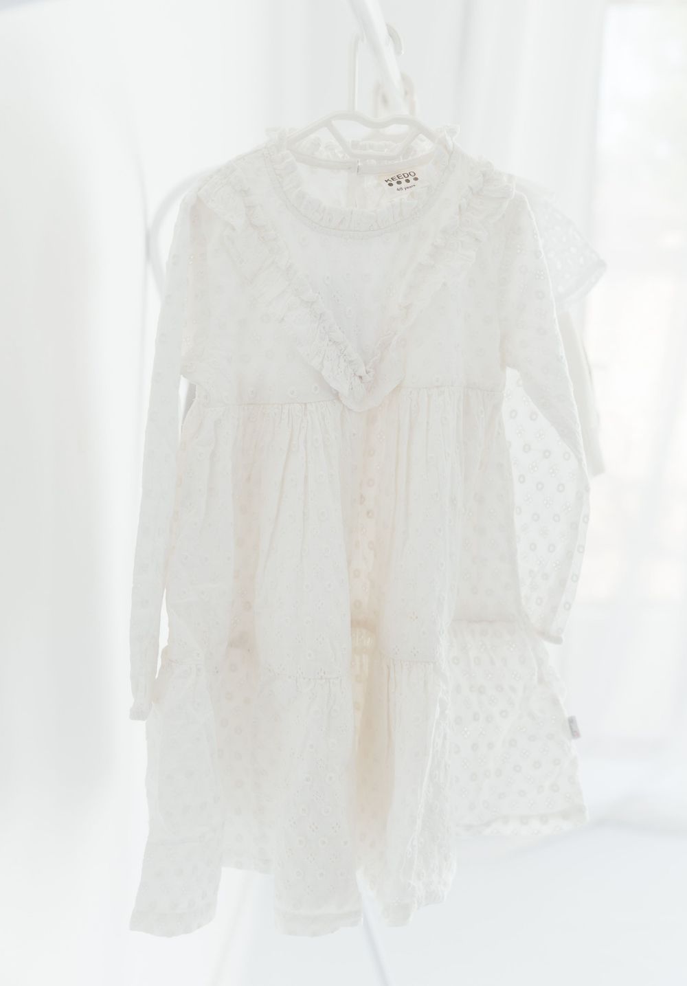 A delicate white lace dress with long sleeves hangs on a hanger against a light background.