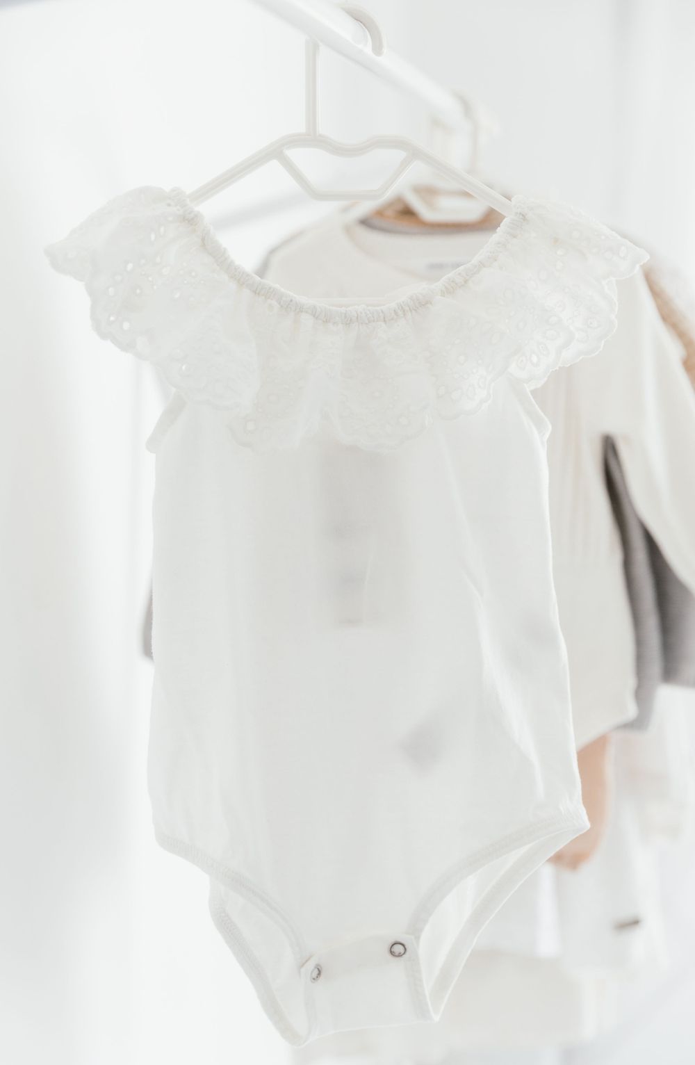 White baby bodysuit with delicate lace ruffle collar hanging on white hanger against light background.