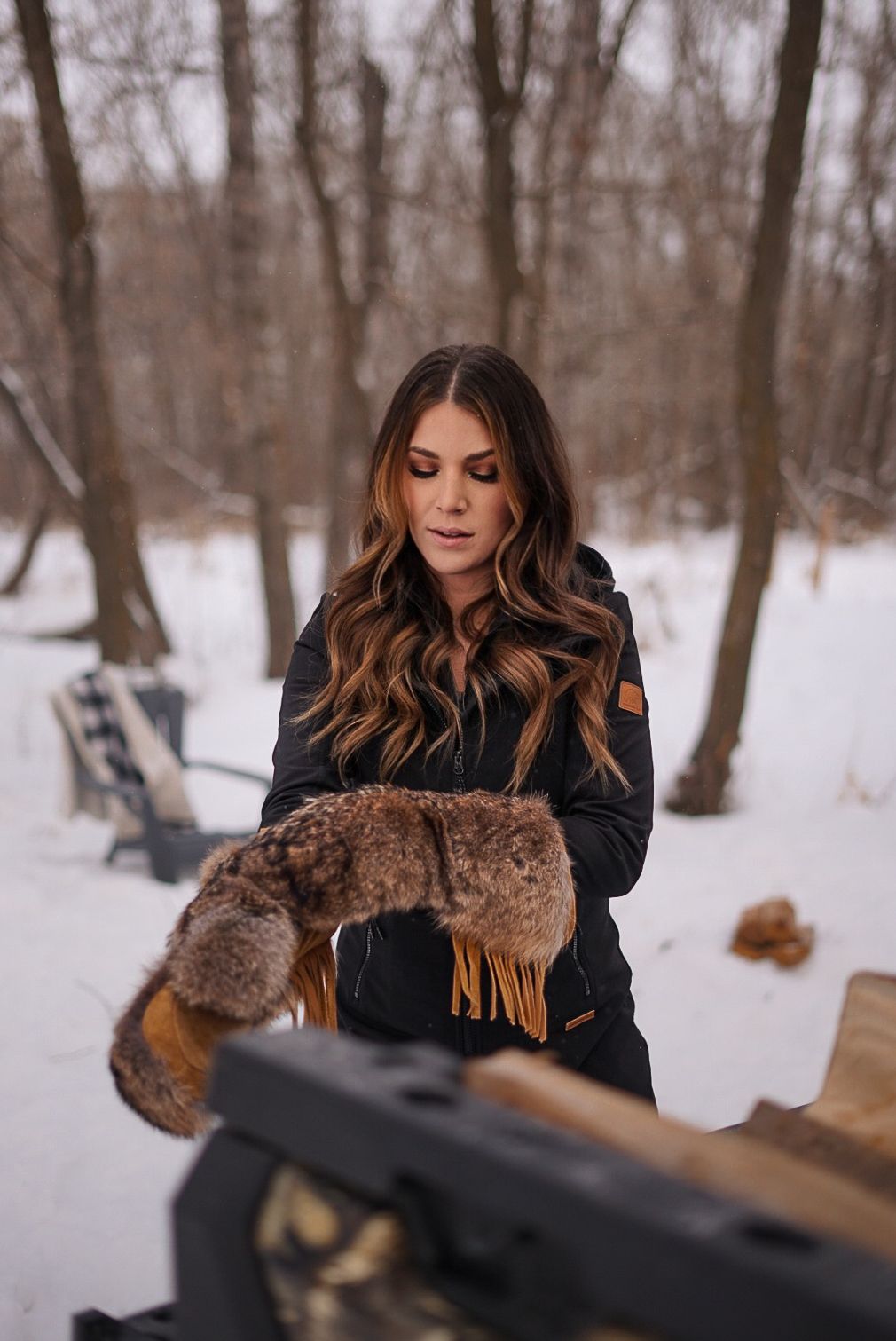 Person wearing black winter coat holding fur accessories in snowy forest setting.