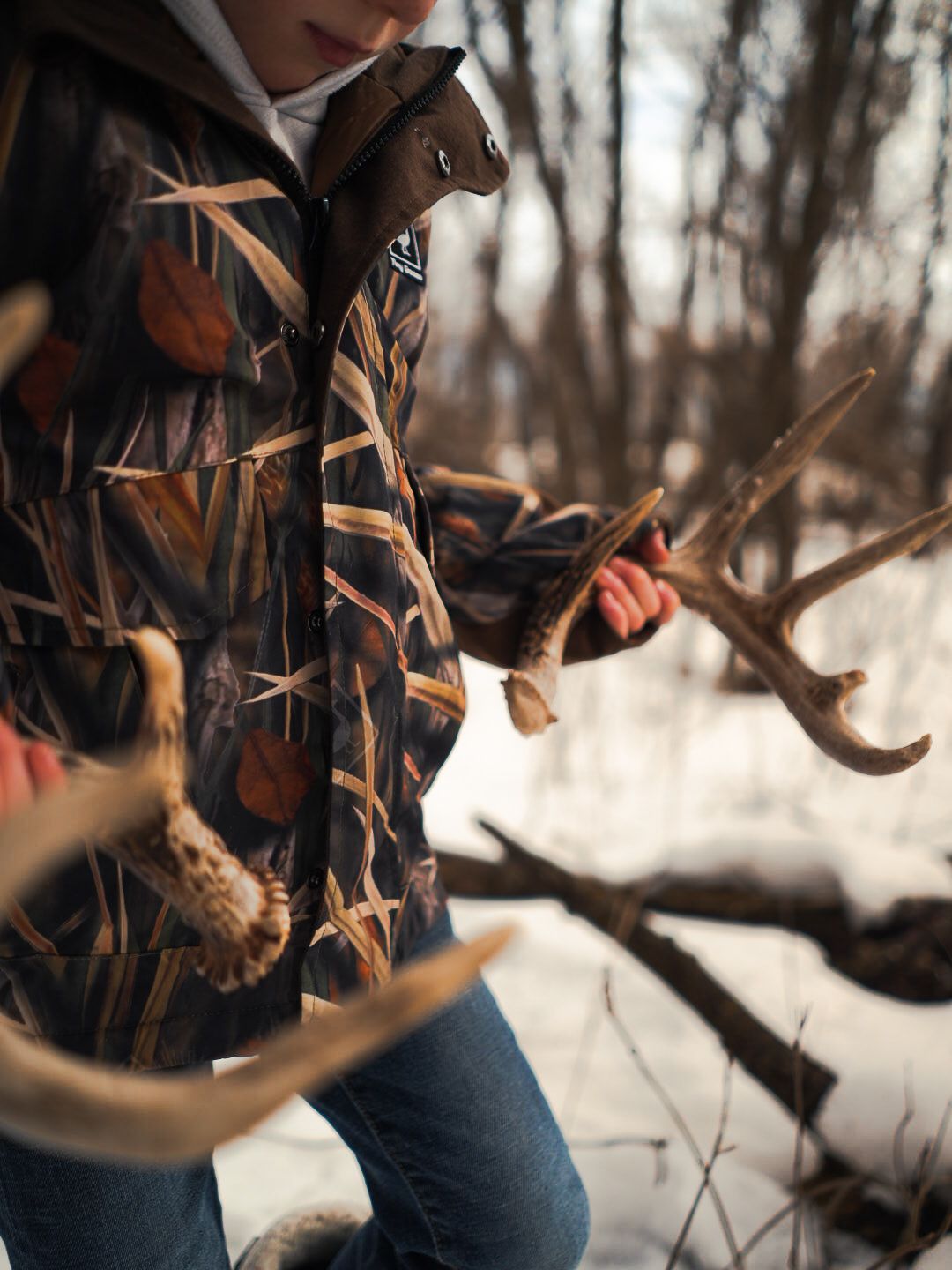 Close-up of camouflage jacket and deer antlers in winter forest setting.
