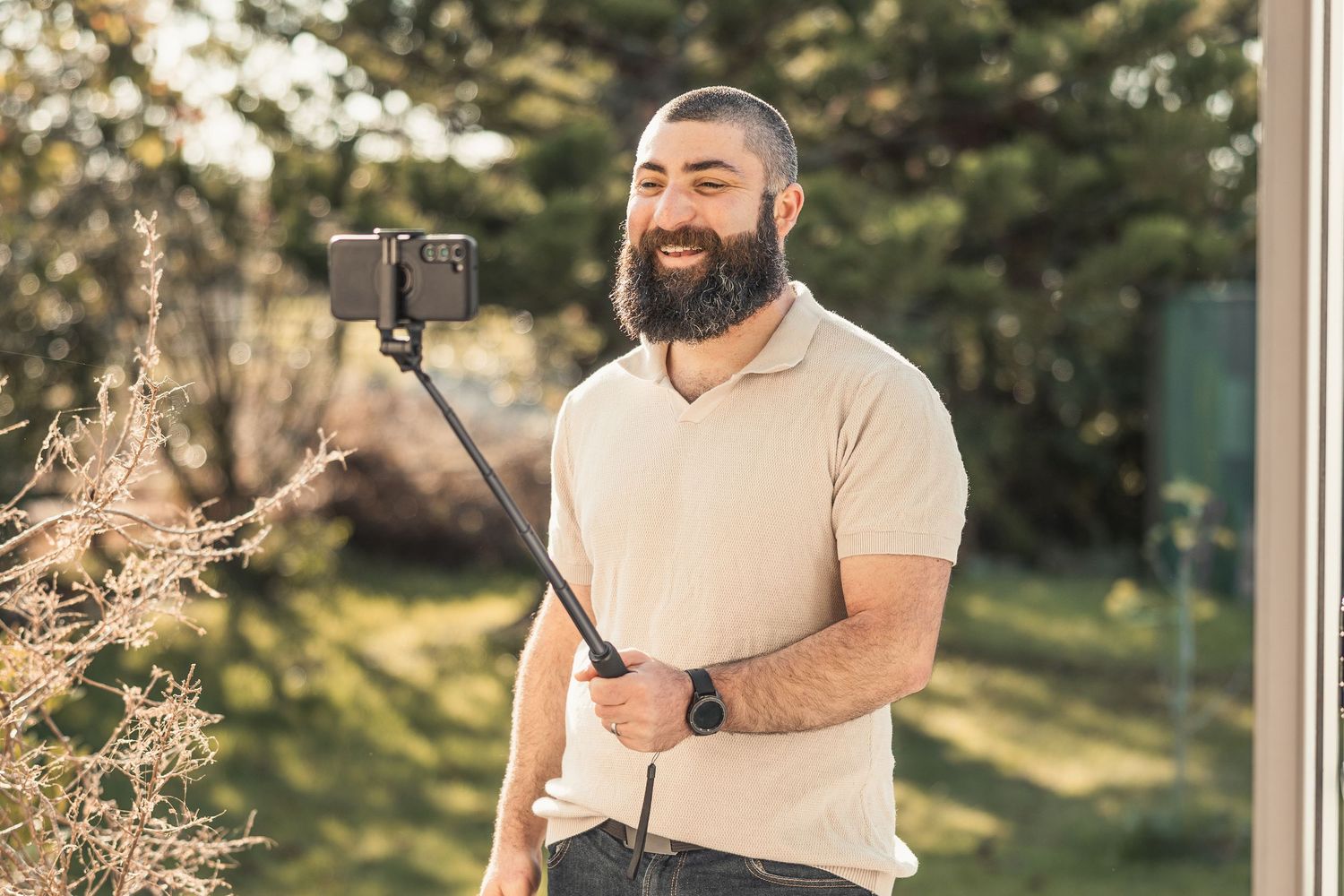 A person with a beard wearing a light polo shirt holds a selfie stick while recording outdoors in a sunny garden.