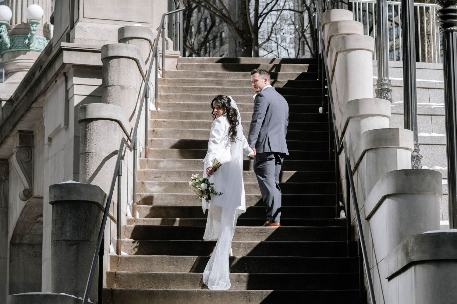 Couple dancing in a dramatic dip pose on a modern covered bridge walkway with metal railings and city backdrop.