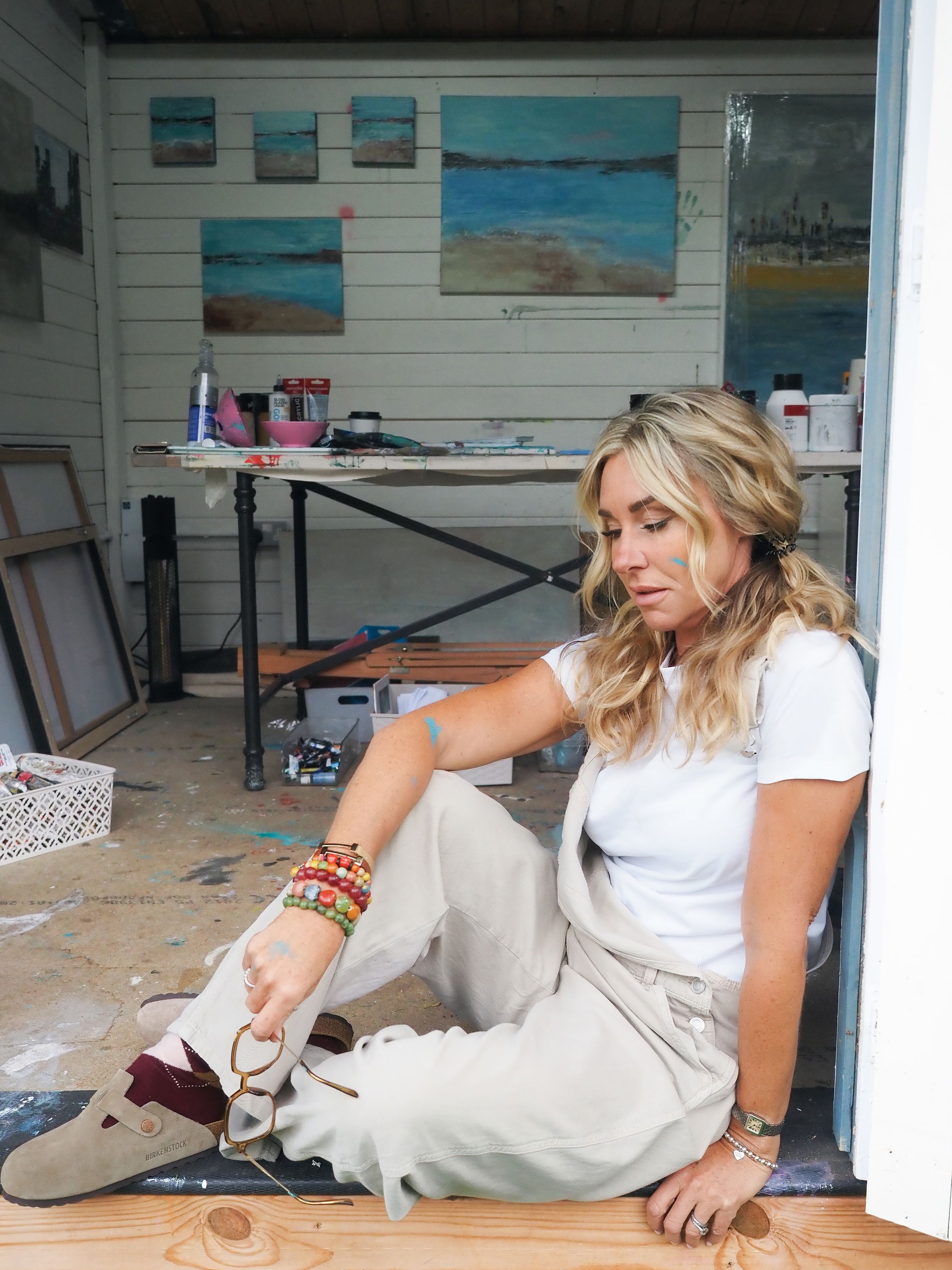 Artist sits on floor of studio space with colorful beach paintings displayed on white walls in background.