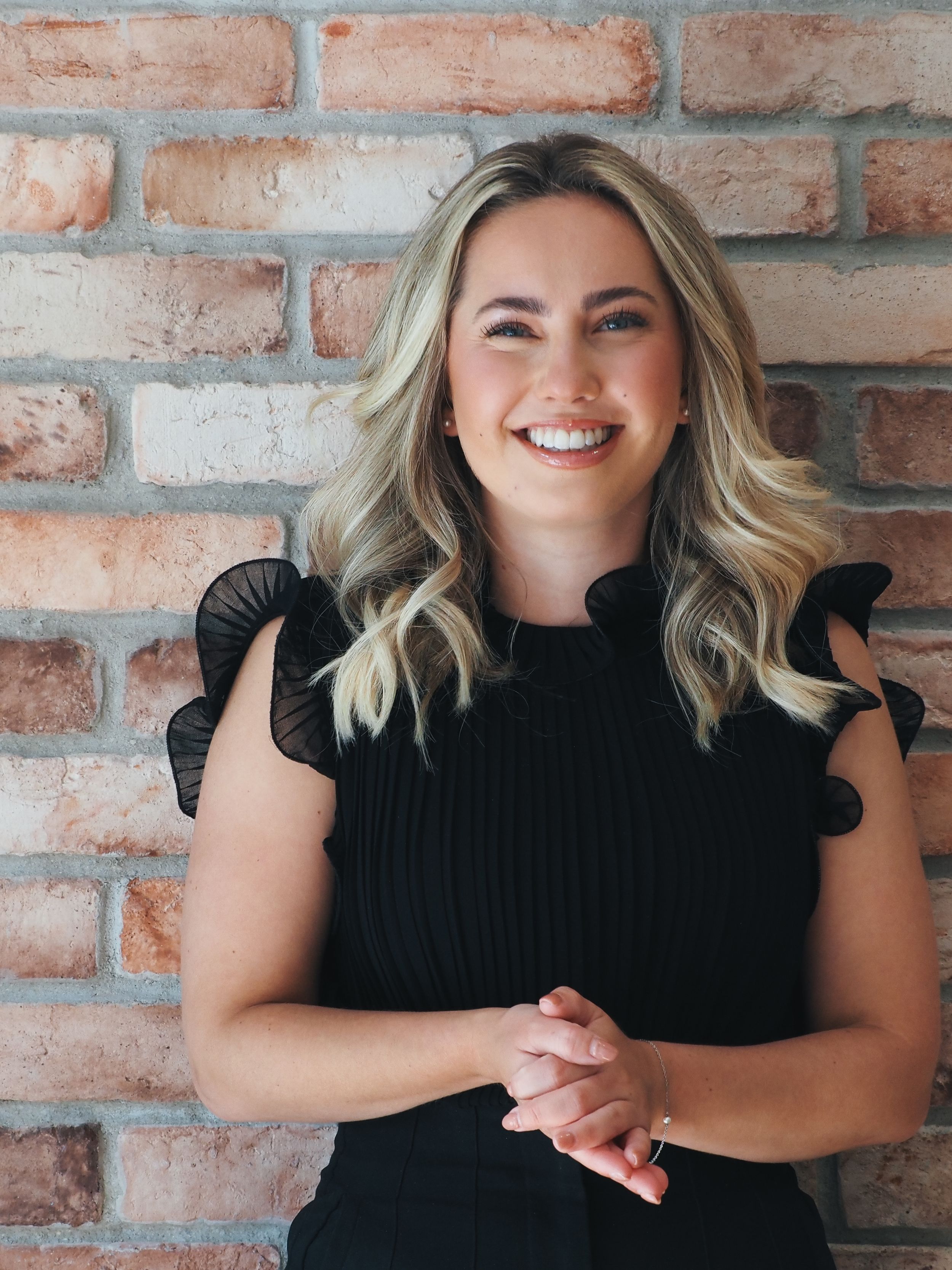 Professional portrait of a woman in a black top with flutter sleeves standing against a rustic brick wall background.