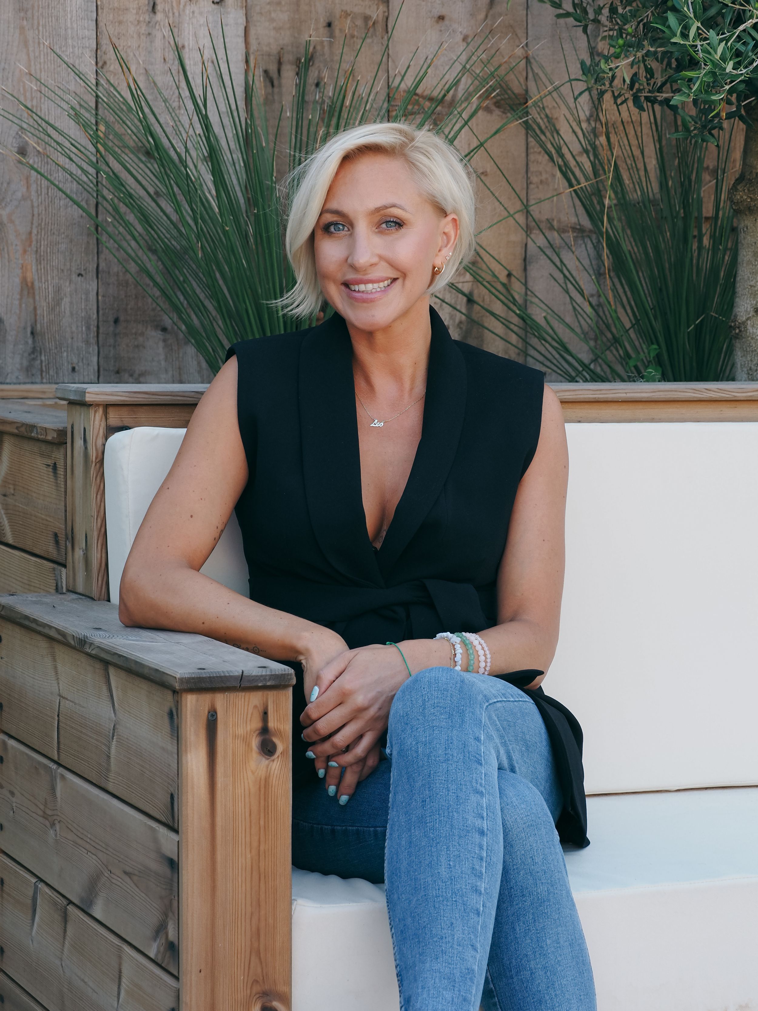 Woman in black sleeveless top and jeans sits on wooden bench with greenery in background.