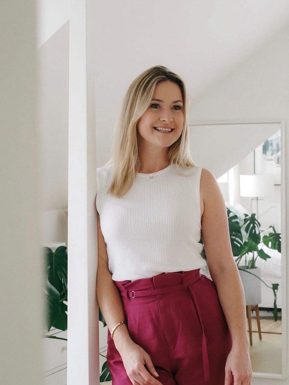 Woman in white sleeveless top and burgundy high-waisted pants poses in a bright room with houseplants.
