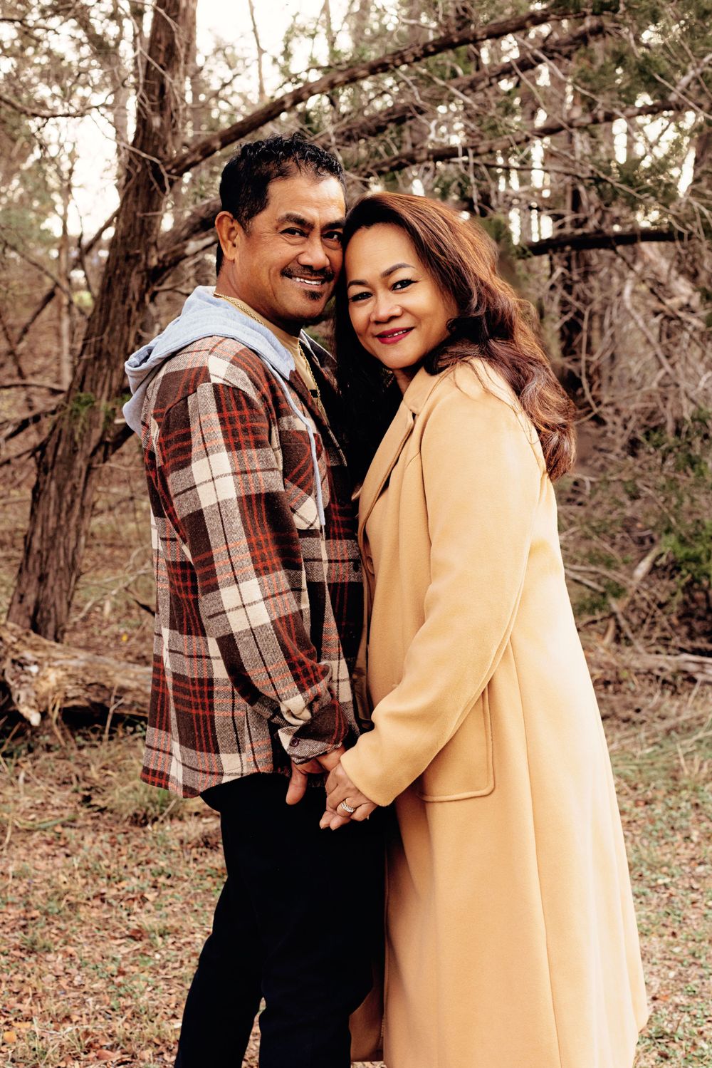 A couple poses together outdoors in autumn, wearing a plaid shirt and yellow coat while smiling at the camera.