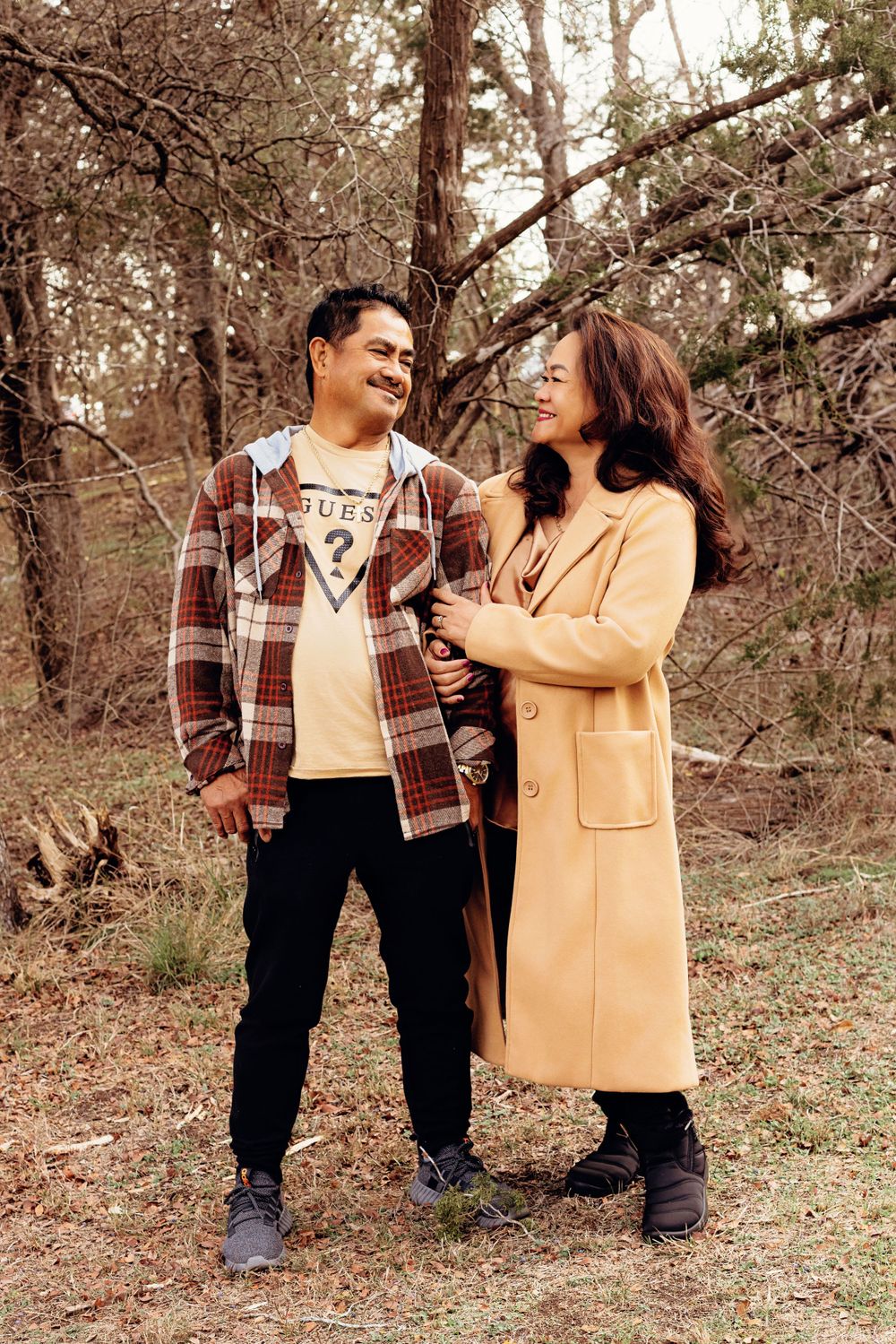 A happy couple laughs together while posing in a forest setting wearing coordinated casual outfits.