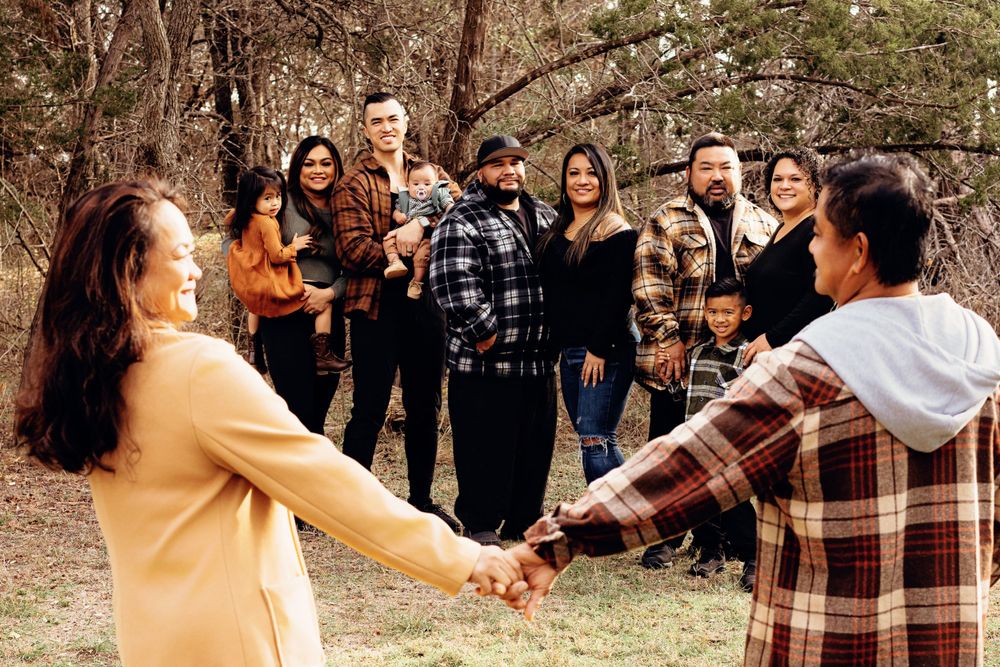 A group of people holding hands in a circle outdoors during autumn surrounded by trees and fallen leaves.
