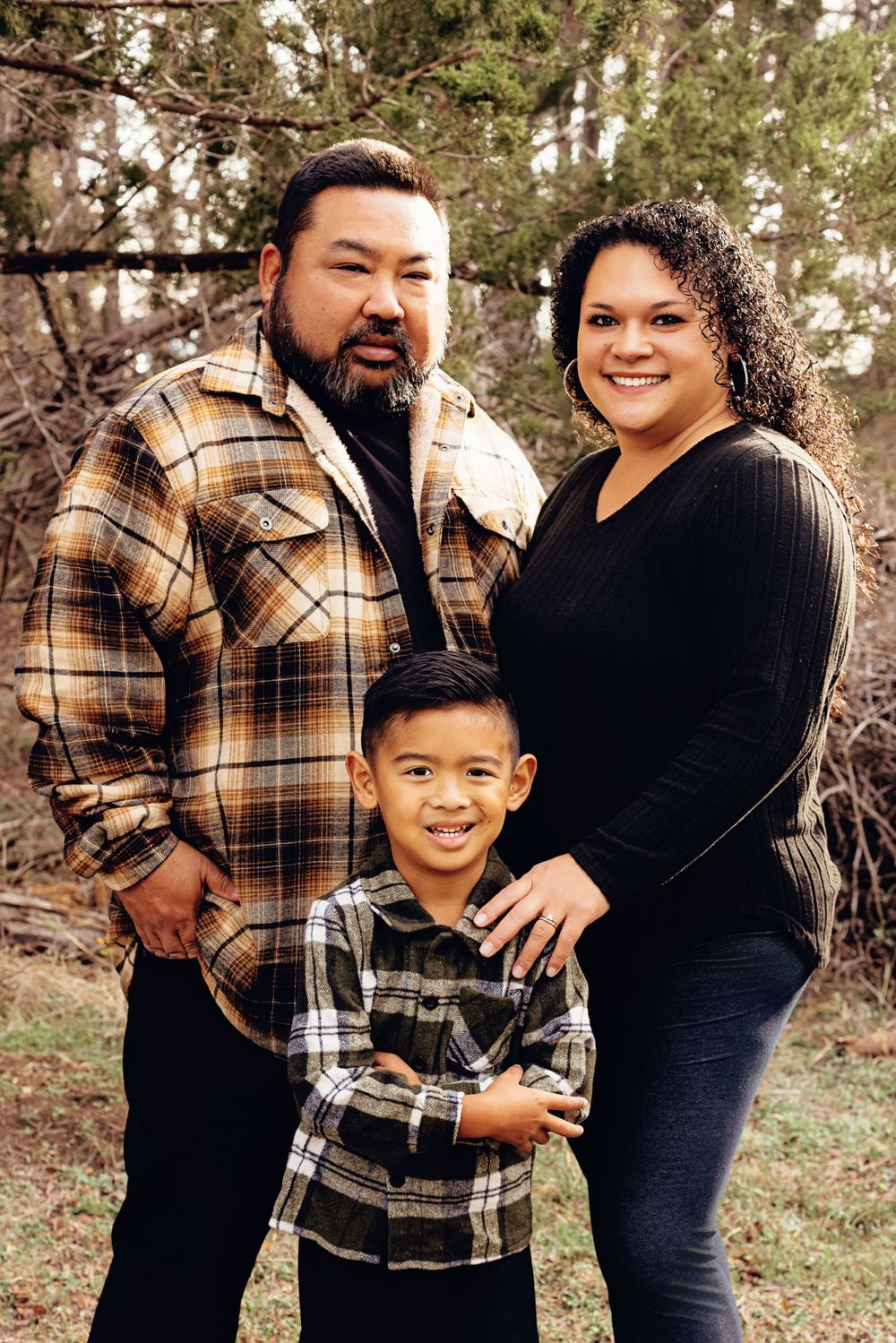 A family poses together outdoors in plaid shirts and dark pants against a natural wooded backdrop.