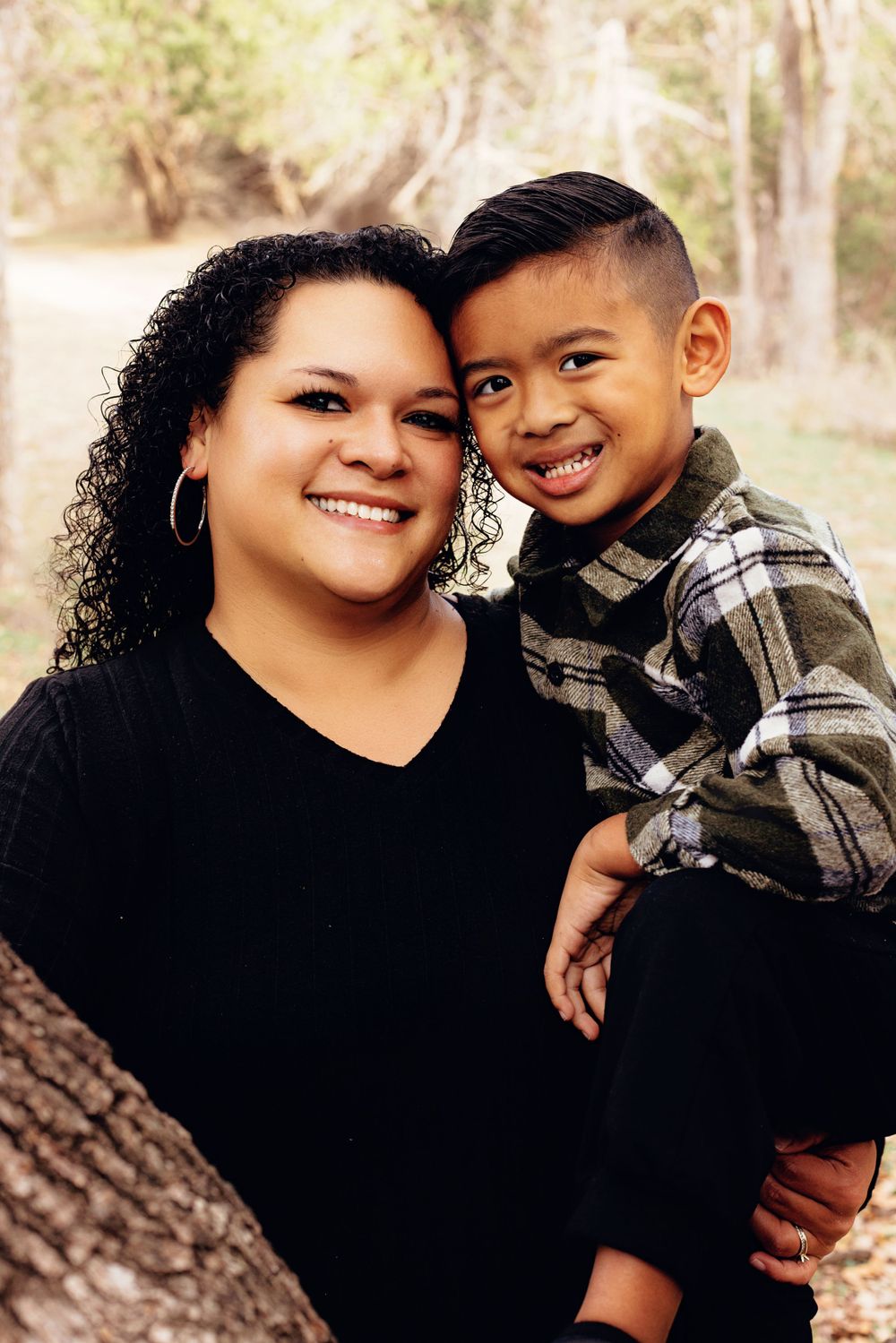A parent and child pose together outdoors, both smiling warmly while wearing casual clothing.