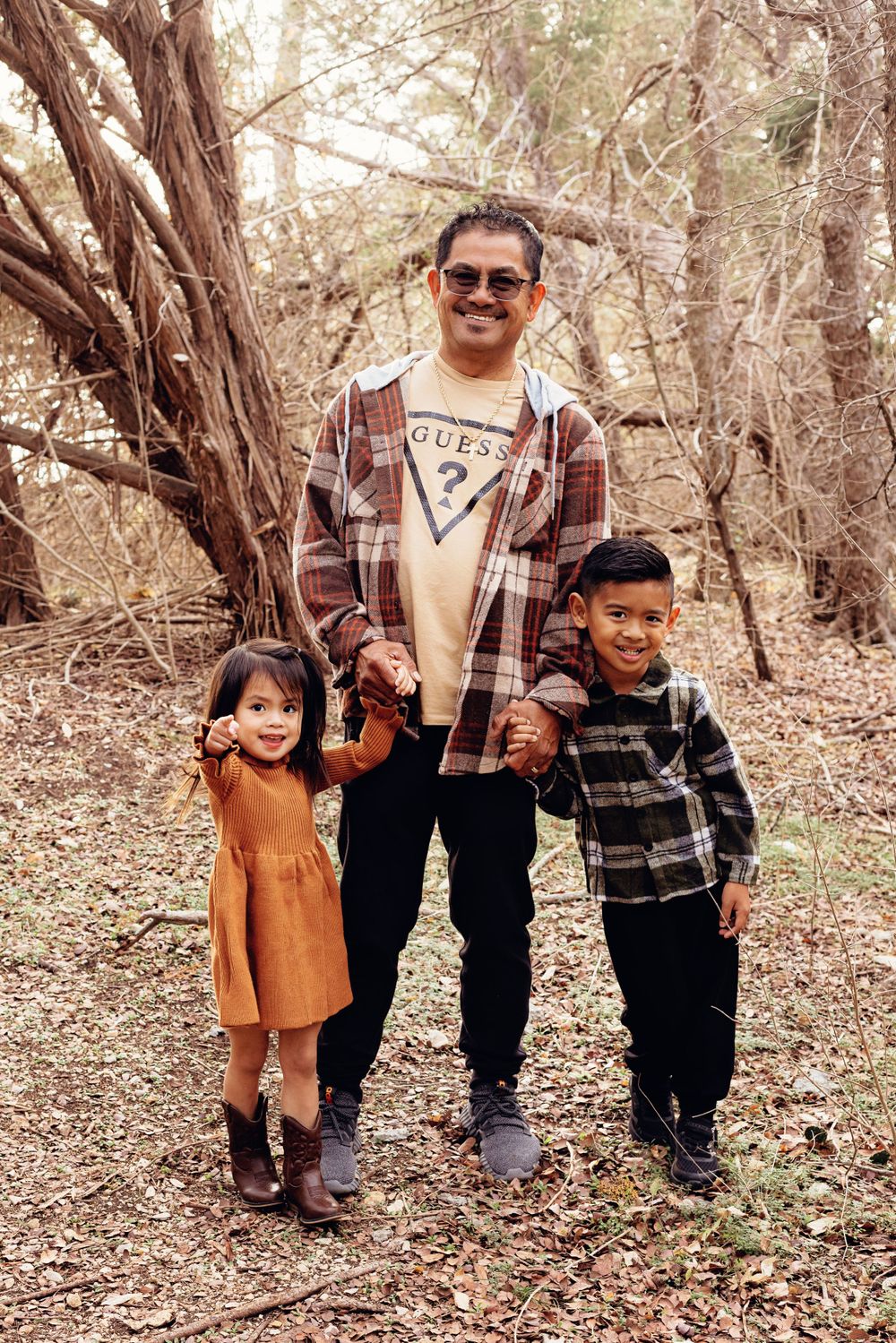 Three people stand together outdoors in a wooded area wearing plaid and casual clothing.