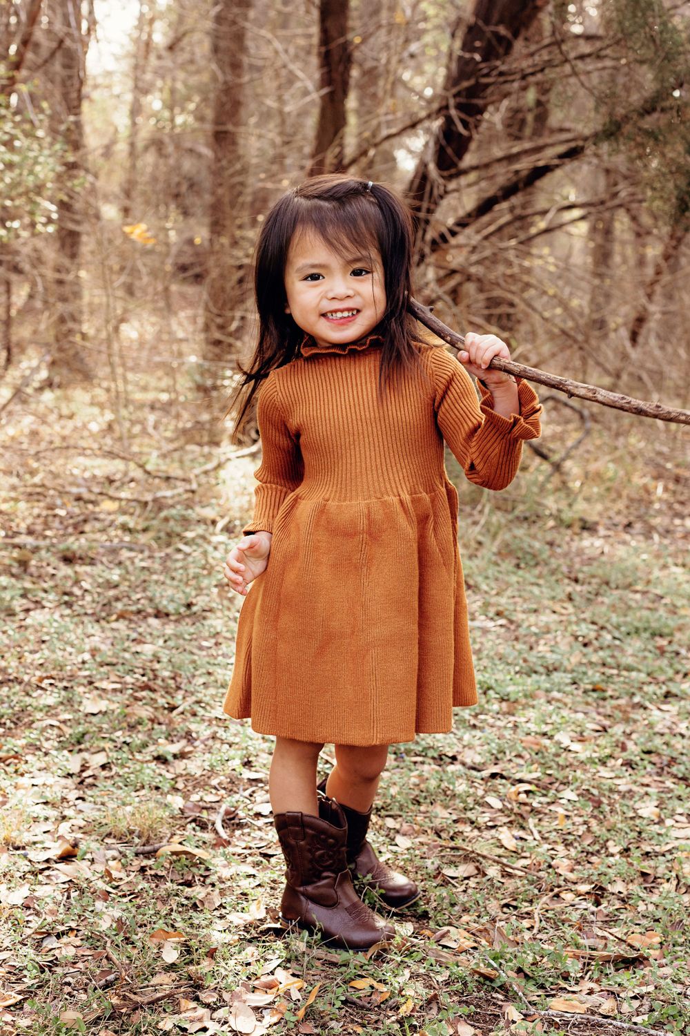 A small child in a rust-colored dress smiles while playing in a wooded area.