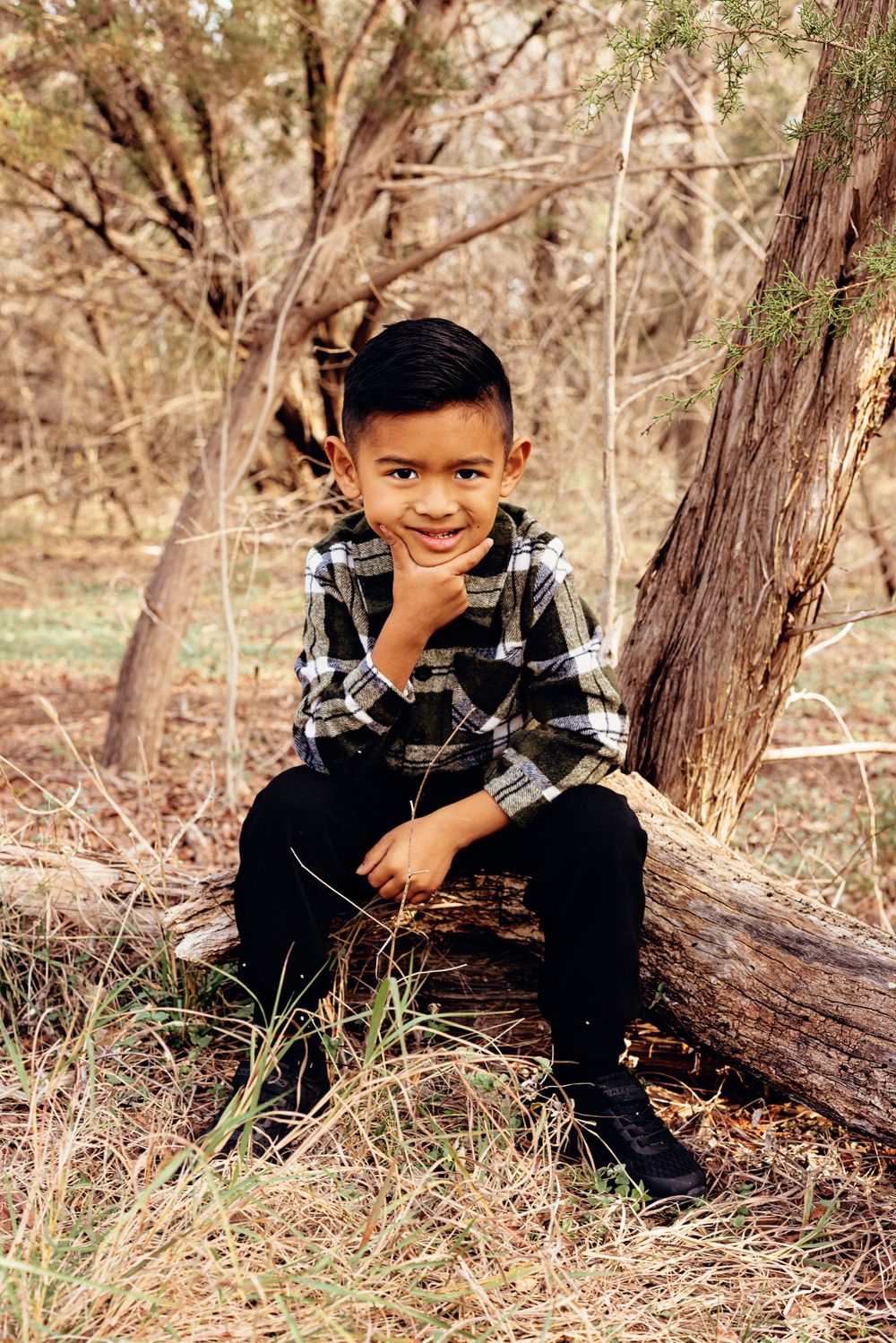Child in plaid flannel shirt and black pants sits thoughtfully on fallen log in autumn woods.
