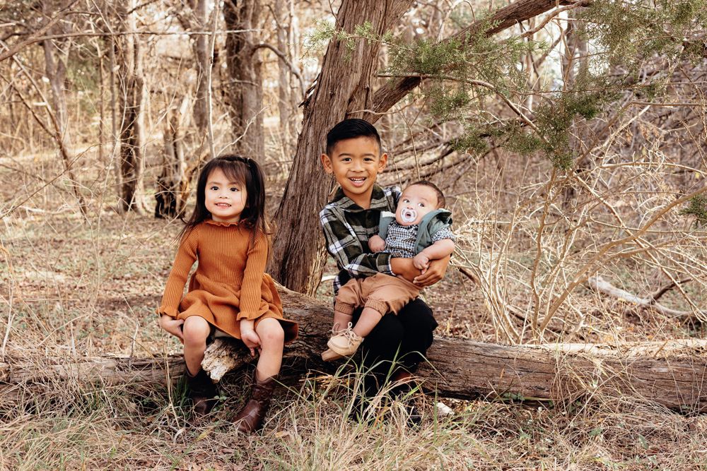 Two children sit together on a fallen log in an autumn forest wearing cozy sweaters and boots.