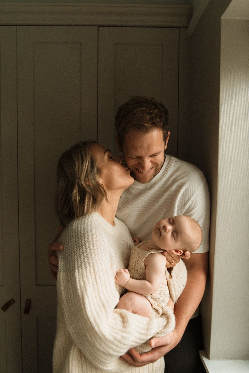 A tender family moment as parents embrace their newborn baby in a softly lit room with white walls.