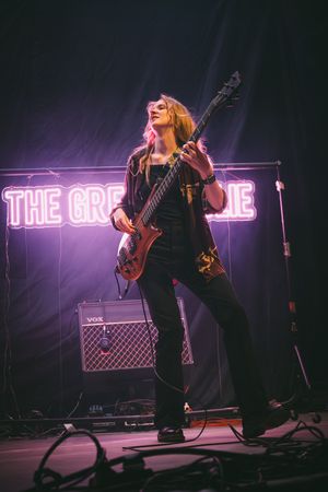 Female guitarist performing on stage at The Greenie venue, playing bass guitar under purple neon lights.