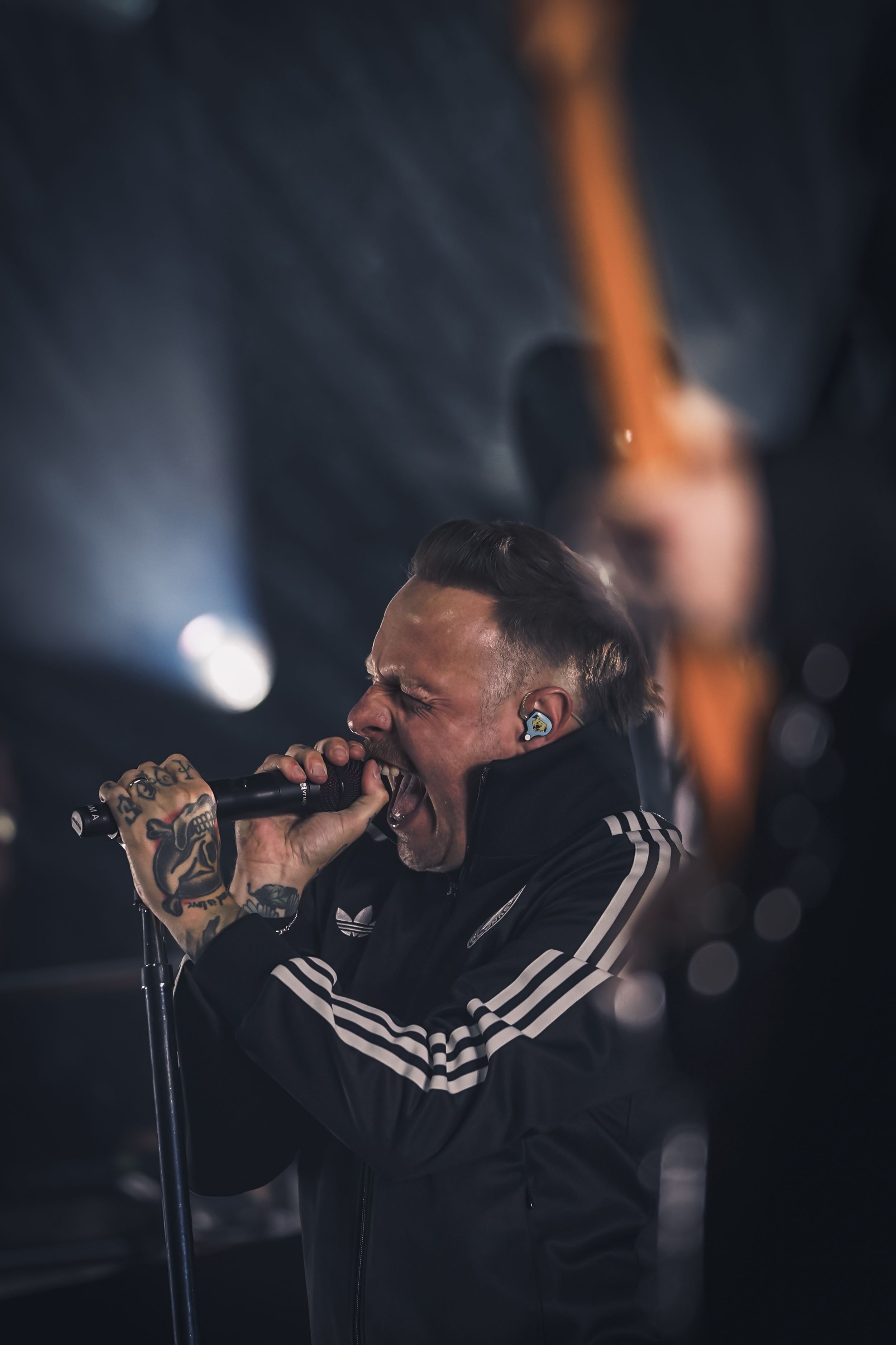 A performer in an Adidas track jacket sings passionately on stage with dramatic lighting in a dark concert setting.