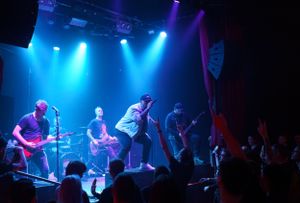 Concert stage bathed in blue lighting with performers silhouetted against dramatic spotlights during a live show.