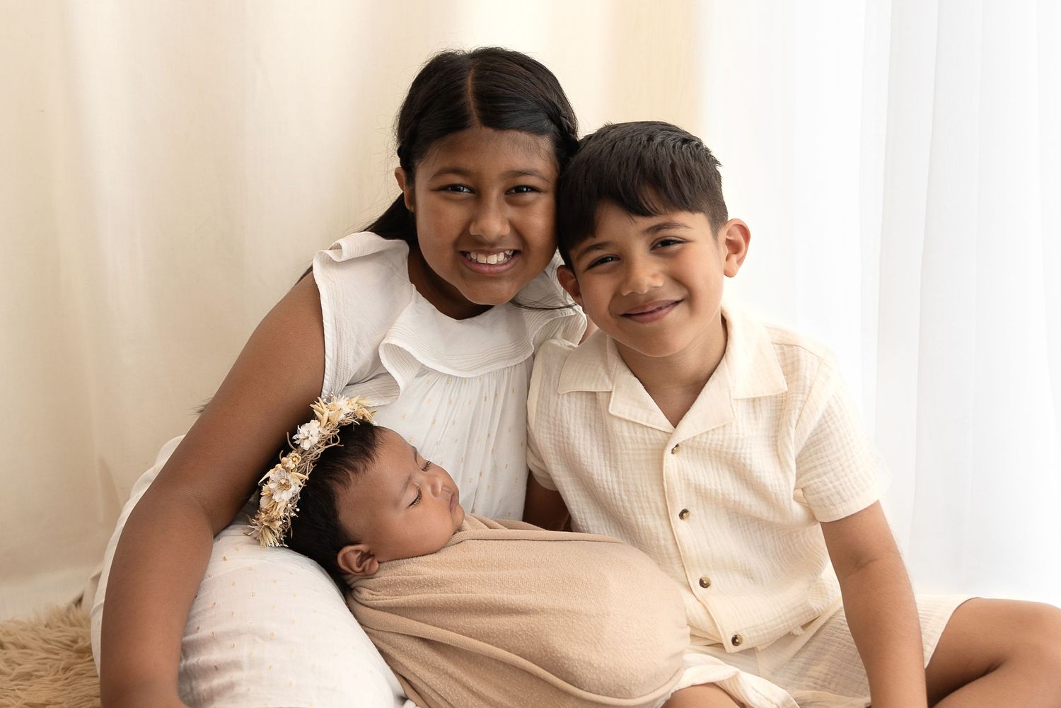 Three children sit together in a tender family moment wearing light-colored clothing against a neutral background.