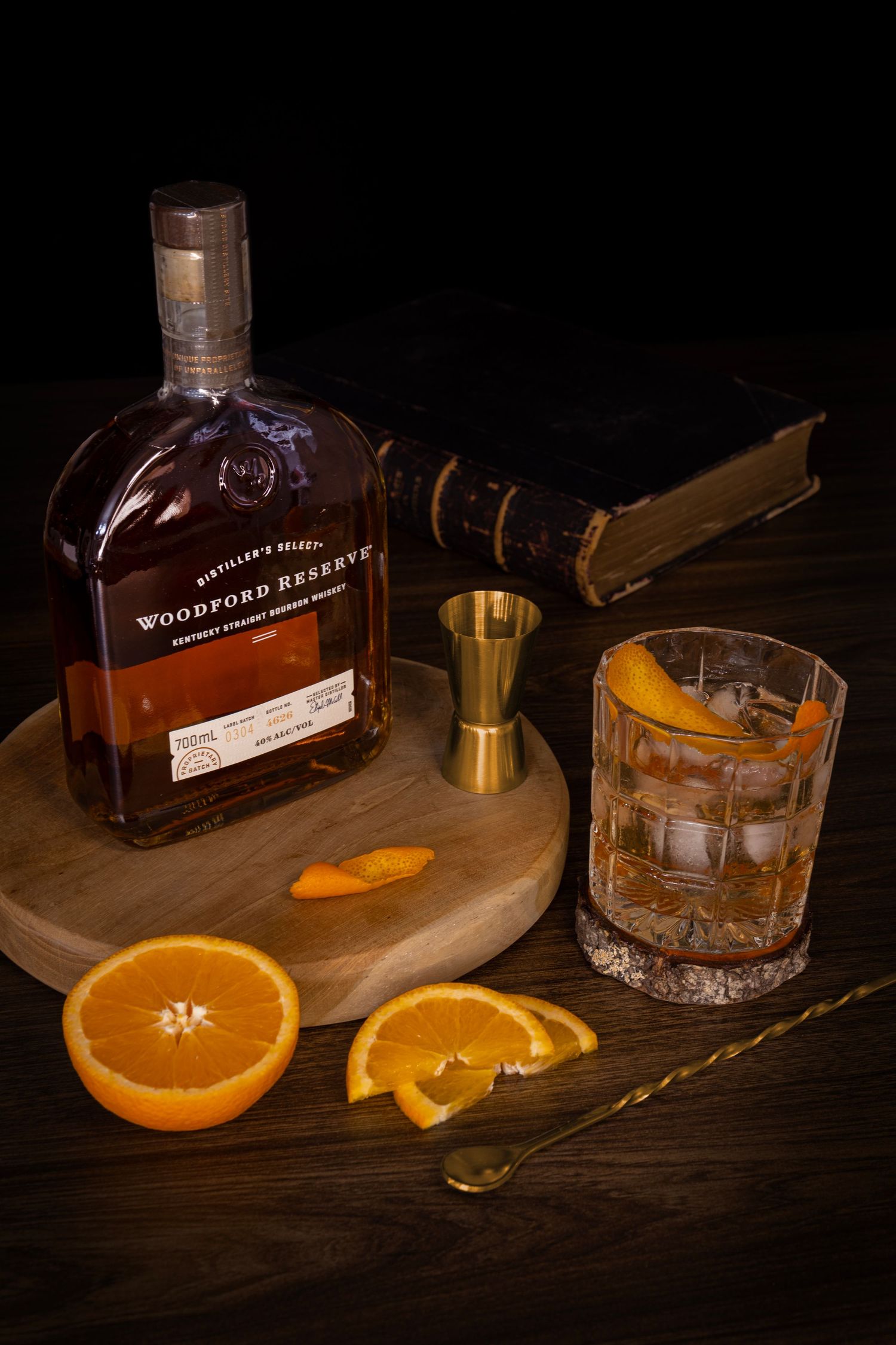 Old Fashioned cocktail with orange twist beside bourbon bottle, orange slices, jigger, and bar spoon on wooden surface.