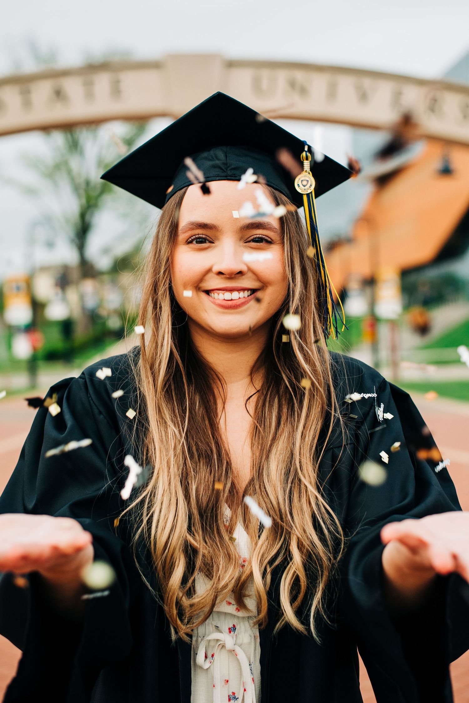 Graduate in black cap and gown smiling outdoors with confetti raining down during commencement celebration.