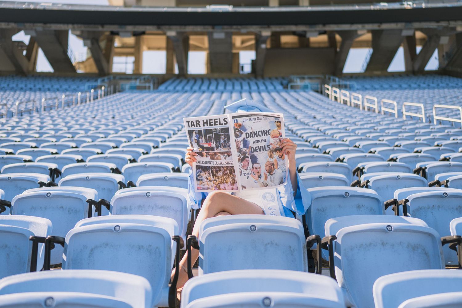 Person reading a book alone in empty stadium seats on sunny day.