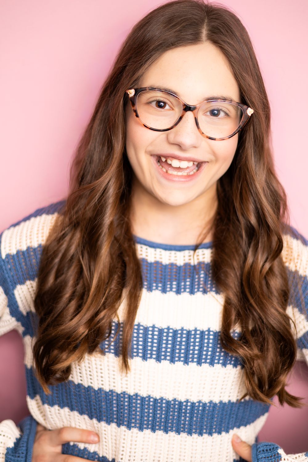 Smiling young girl with long brown hair wearing tortoiseshell glasses and a blue striped knit sweater on pink background.